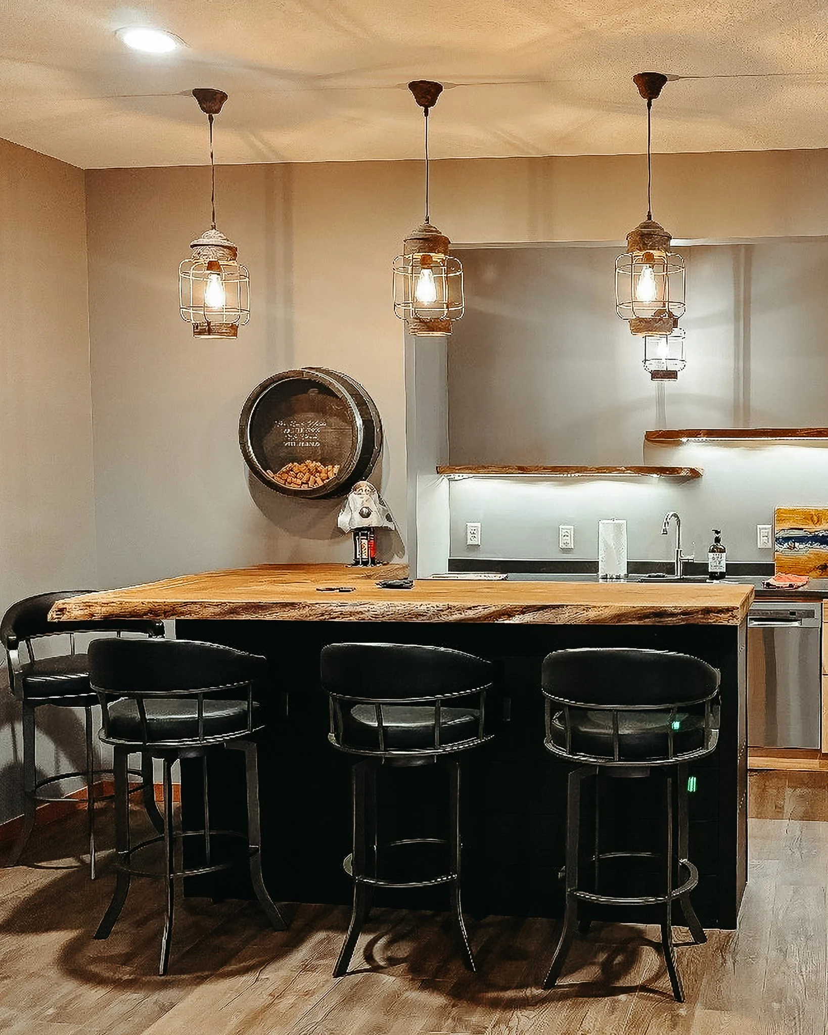 A kitchen or dining area with three hanging pendant lights above a wooden table. Four black chairs are positioned around the table. On the wall, there is a round black cabinet with a reflective surface and a shelf below it. The countertop has a paper