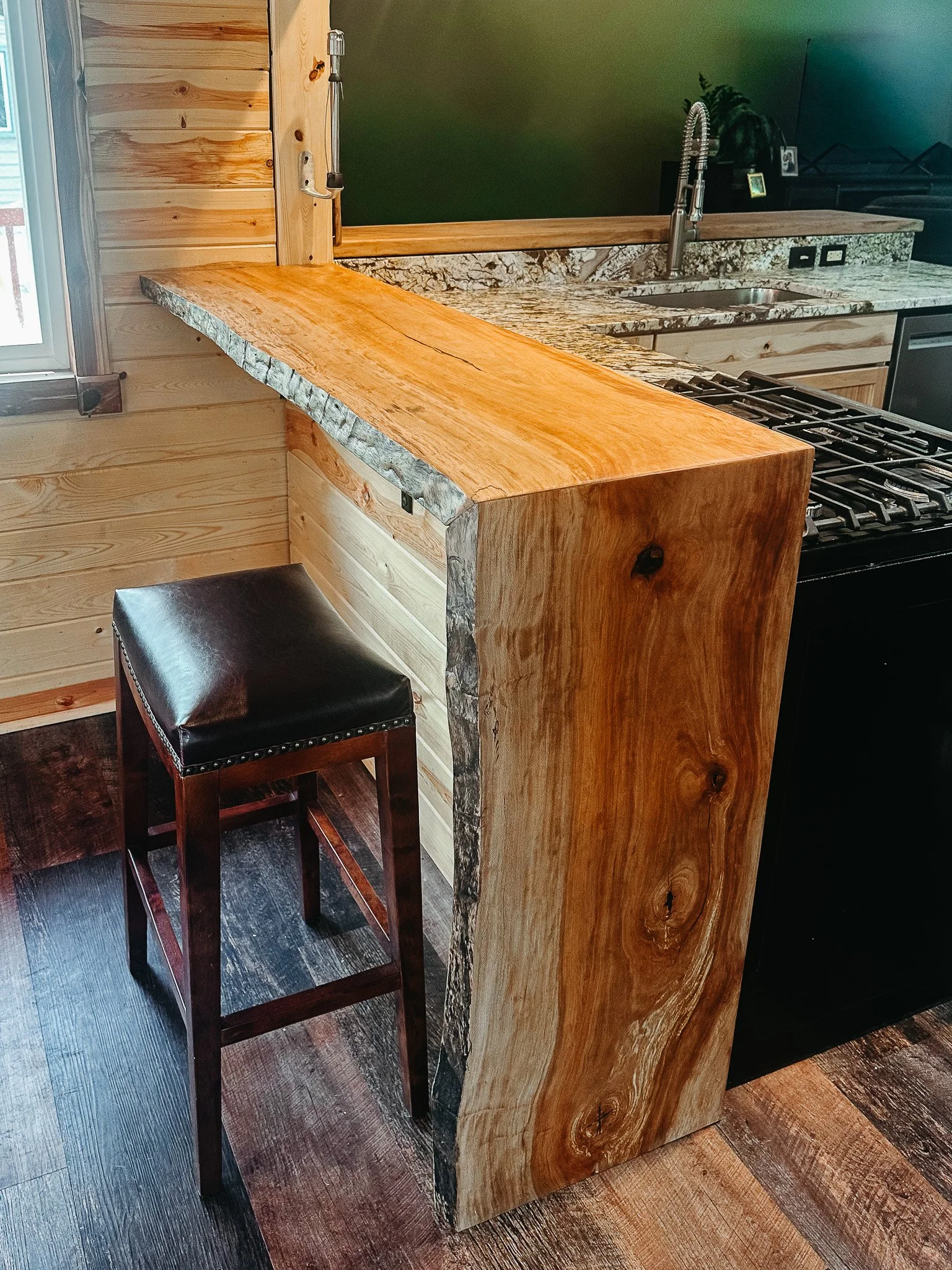 A wooden kitchen counter with a live edge design, showing natural wood grain and knots, positioned next to a smaller matching wooden stool with a black cushioned seat. The kitchen features a granite countertop, a stainless steel faucet, and wooden ca