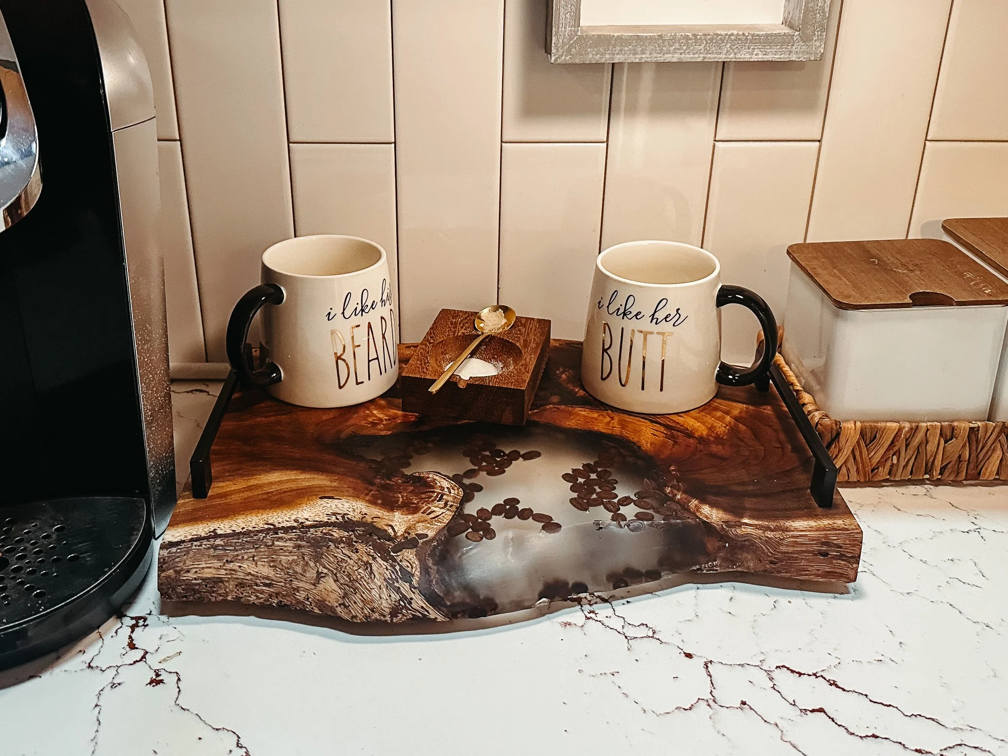 A kitchen countertop with a coffee station featuring two white mugs with black handles, one reading 'I like her BEAR' and the other 'I like her BUT', placed on a wooden tray with a gold spoon. Behind the mugs is a wooden box with a white powder or fl