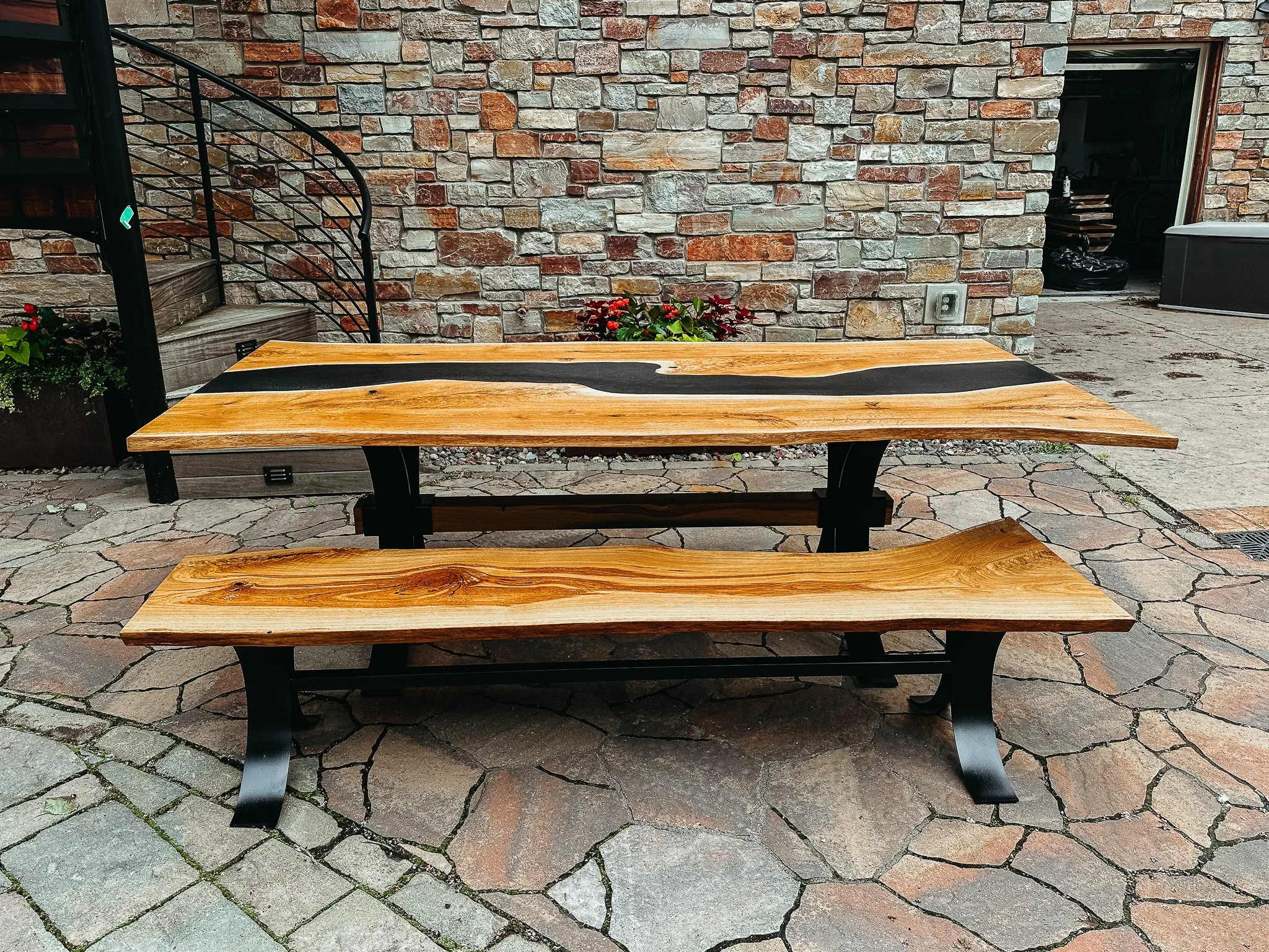 A wooden bench with a black metal frame on a stone patio in front of a brick wall, with some plants and an electrical outlet visible in the background.