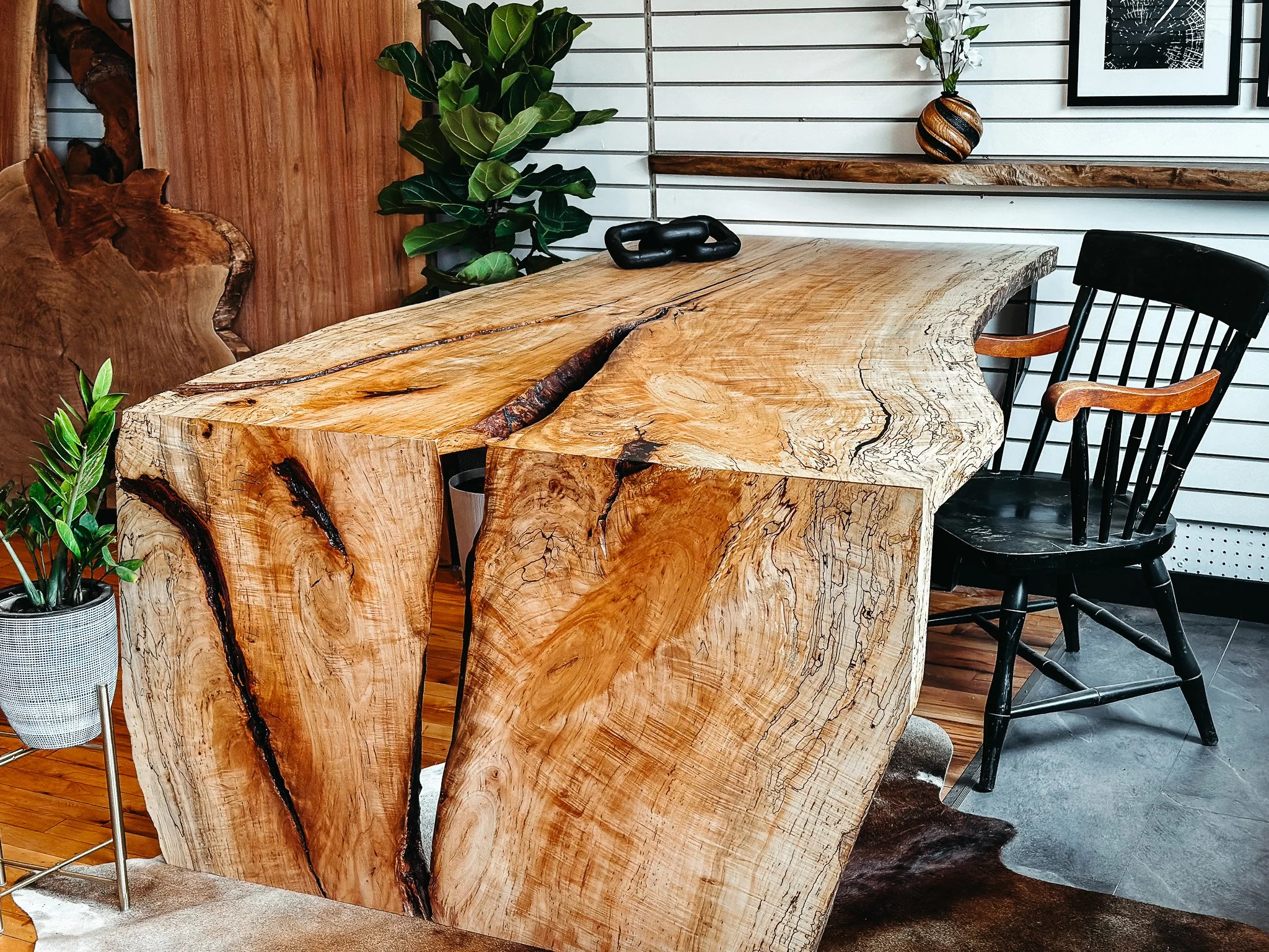 Close-up of a large, natural wood dining table with a live edge in a modern interior space. The table has a textured, polished surface with visible wood grain and cracks.