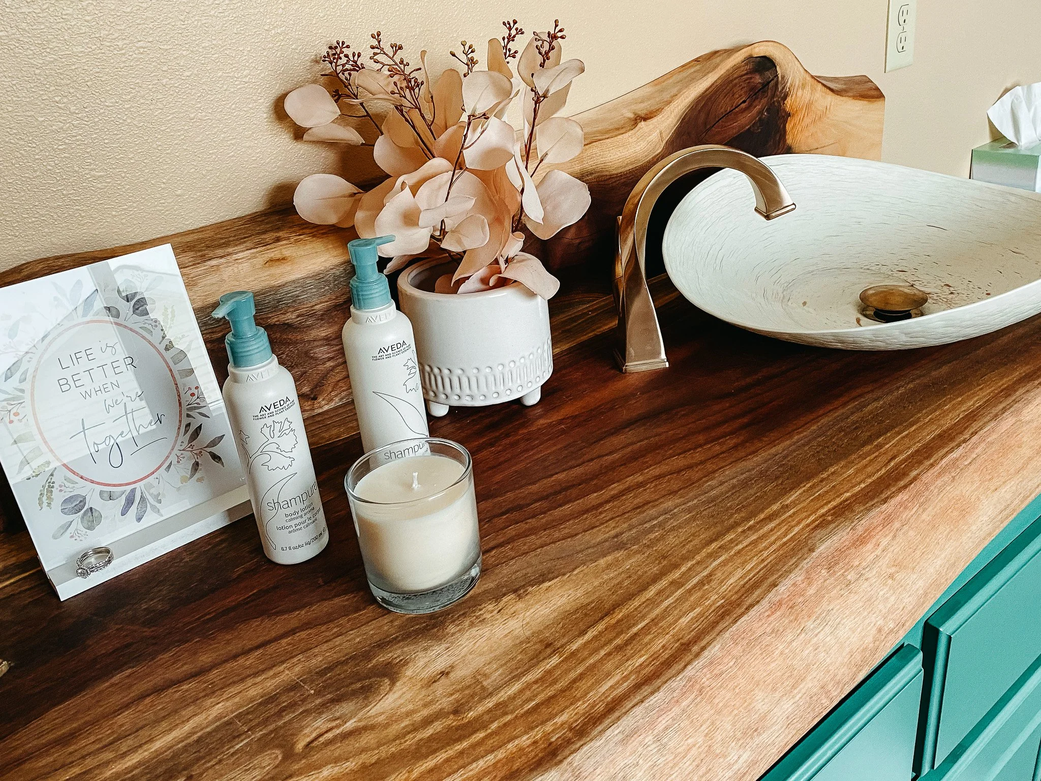 A wooden bathroom vanity with a white vessel sink, a gold faucet, and a decorative wooden piece on the back. There is a vase with pink artificial flowers, three bottles of Avea body lotion, a lit candle, a ring, a sign with the quote "Life is better 