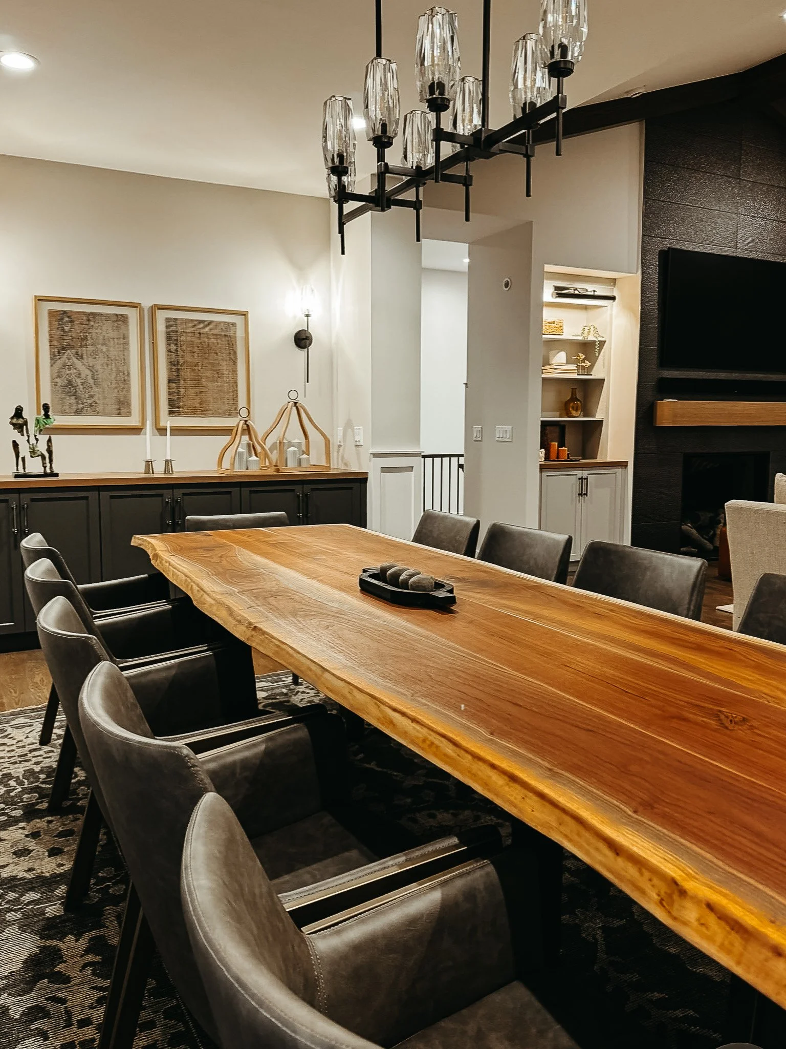 A dining room with a large wooden table surrounded by gray upholstered chairs, decorative artwork on the wall, a black and wood sideboard, and a modern chandelier hanging from the ceiling.