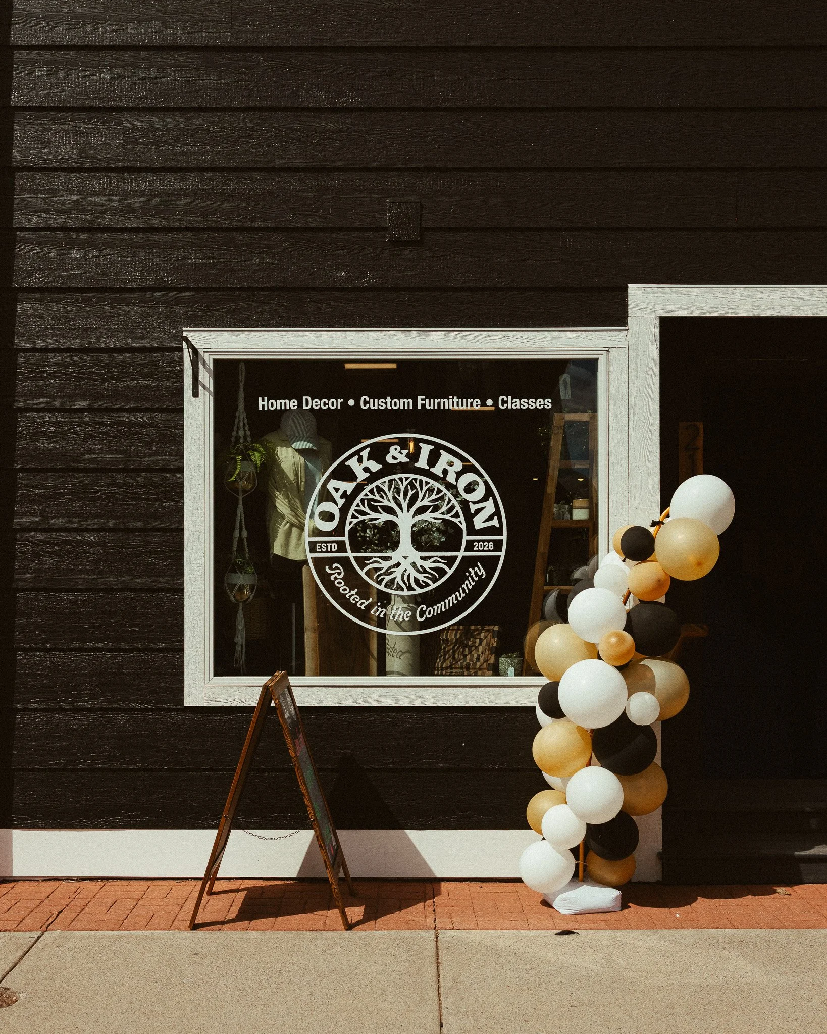 Shop window with white frame, displaying home decor, custom furniture, and classes, with a logo featuring a tree and the text, 'Rooted in the Community.' A cluster of black, white, and beige balloons is outside, along with a standing chalkboard.