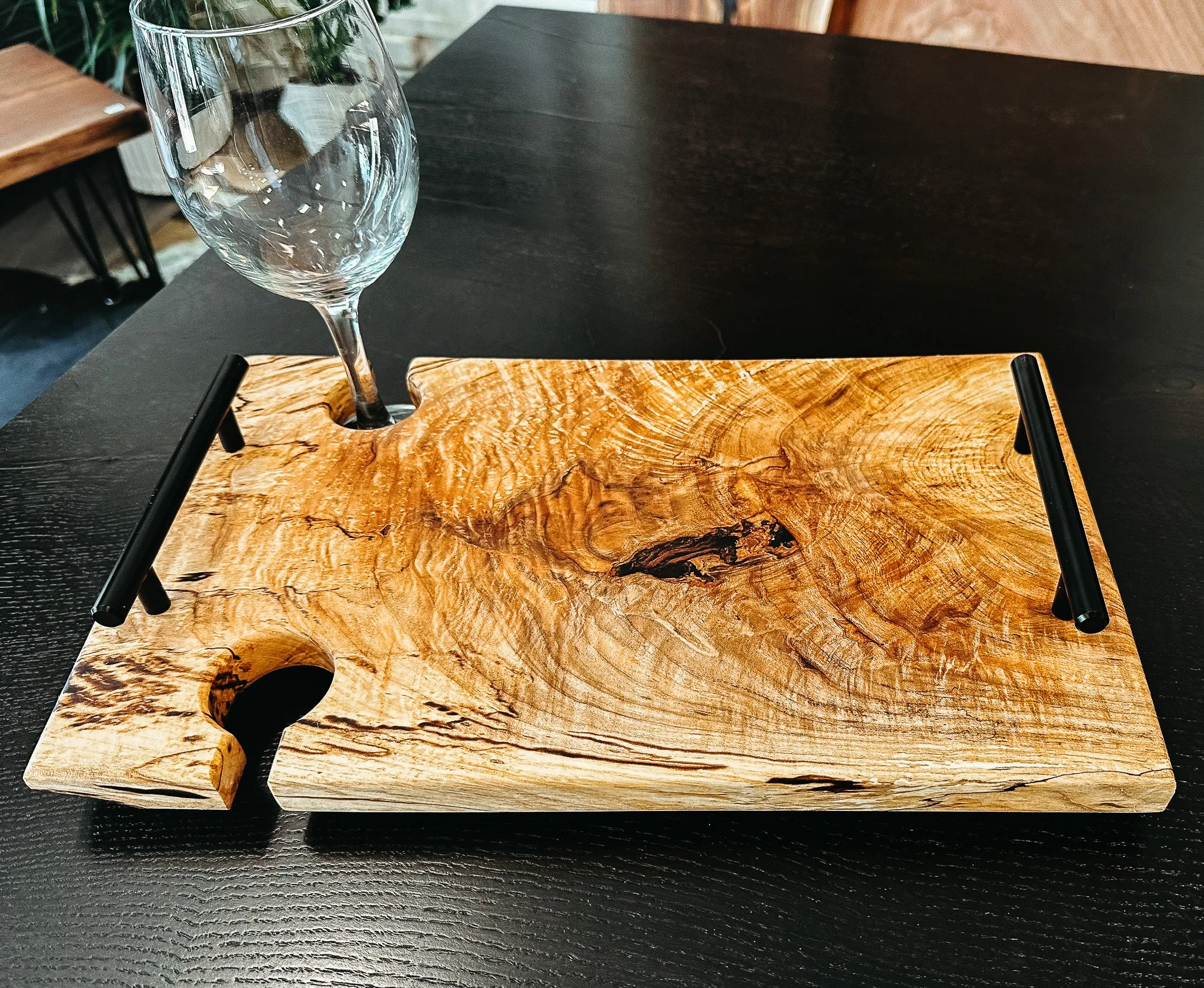 A clear empty wine glass placed on a wooden serving board with black metal handles, on a dark wooden table.