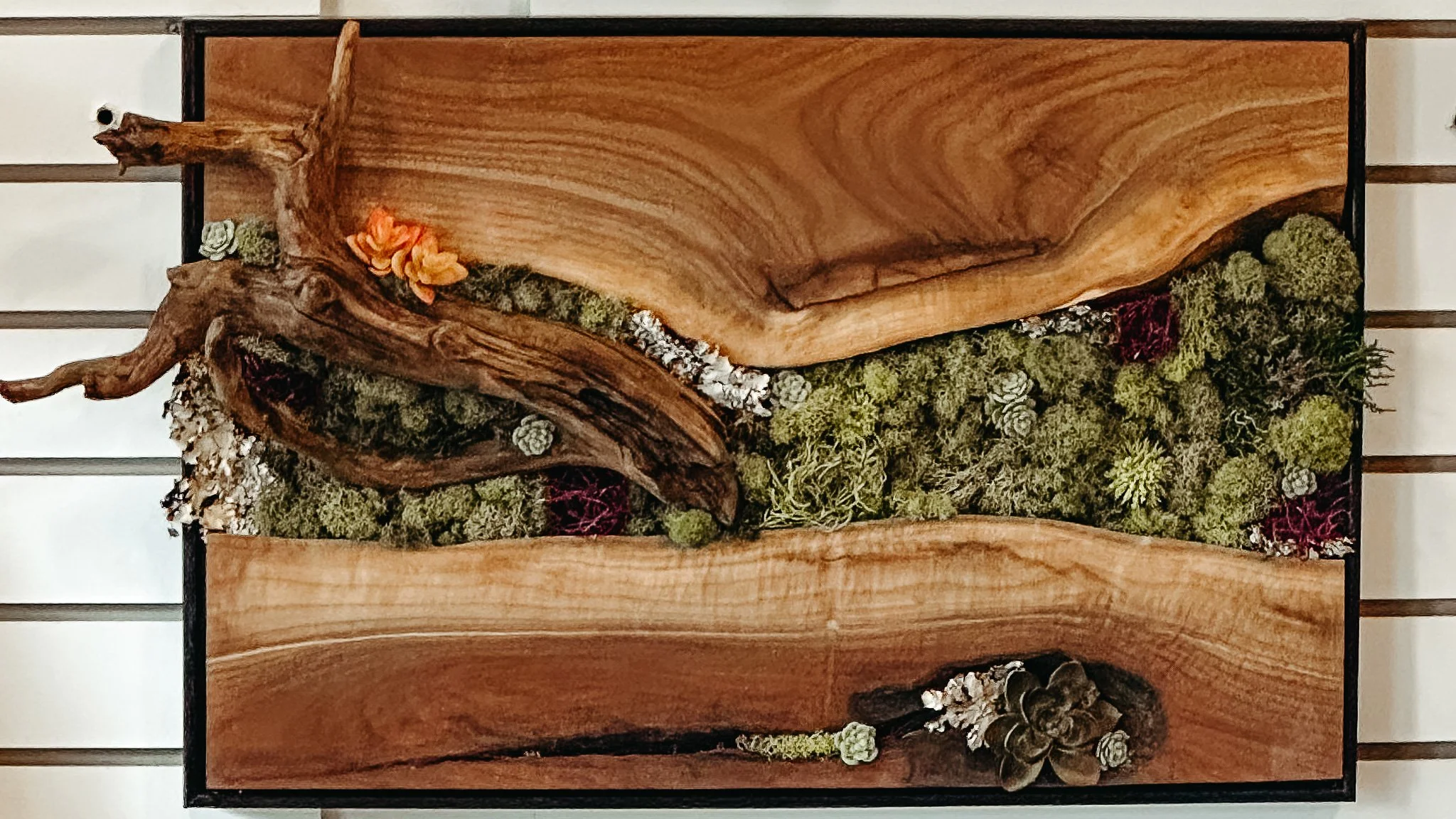 A wooden wall art piece featuring a framed wood panel with natural wood grain, accented with dried moss, pinecones, and small orange and red flowers, arranged artistically.
