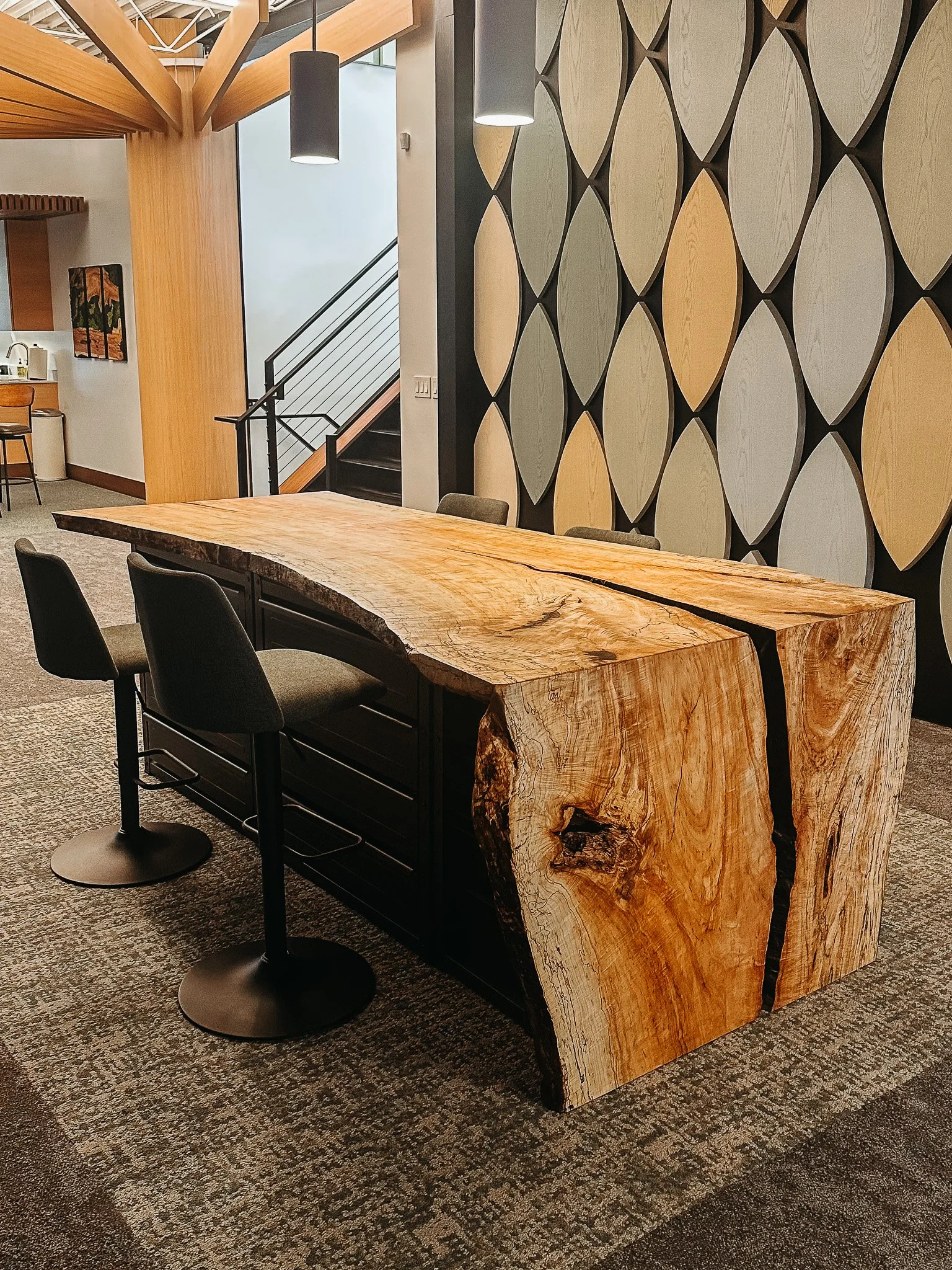 A modern interior space featuring a large, rustic wooden table with natural edges, four chairs, and a wall with a unique circular panel design. There are hanging pendant lights and an abstract painting on a white wall in the background.