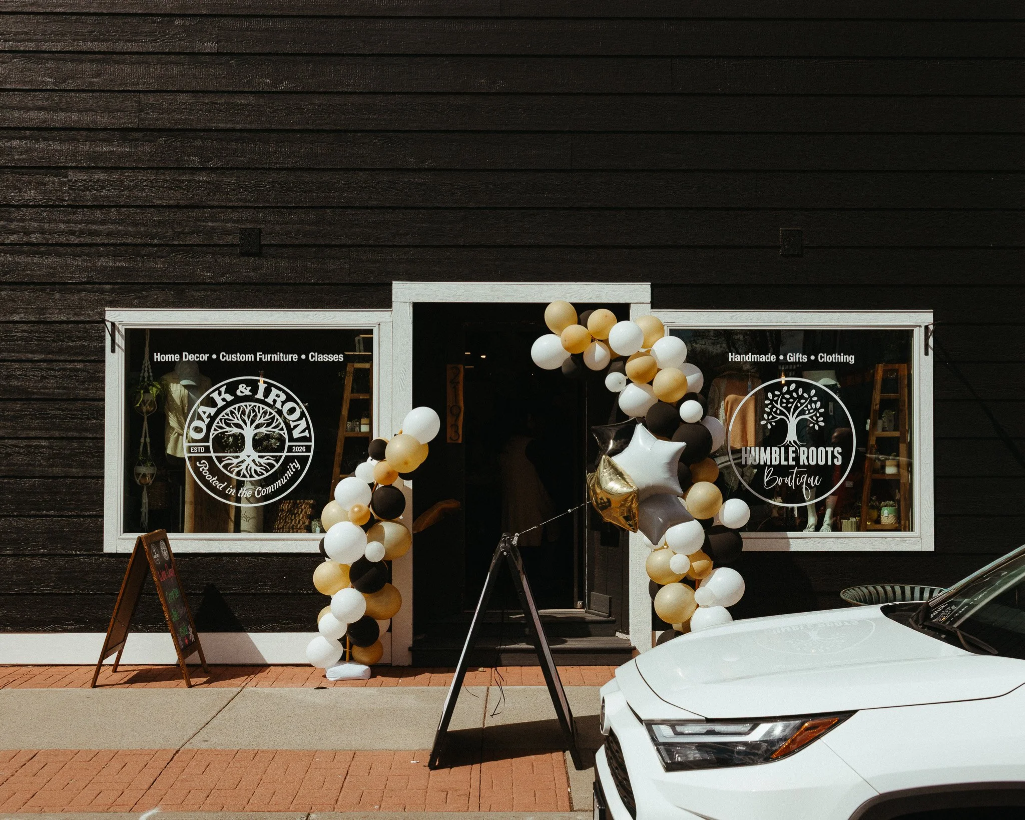 storefront with black wooden exterior, decorated with a balloon arch in black, white, and gold, and featuring a sign for a boutique that sells home decor, custom furniture, handmade gifts, and clothing.