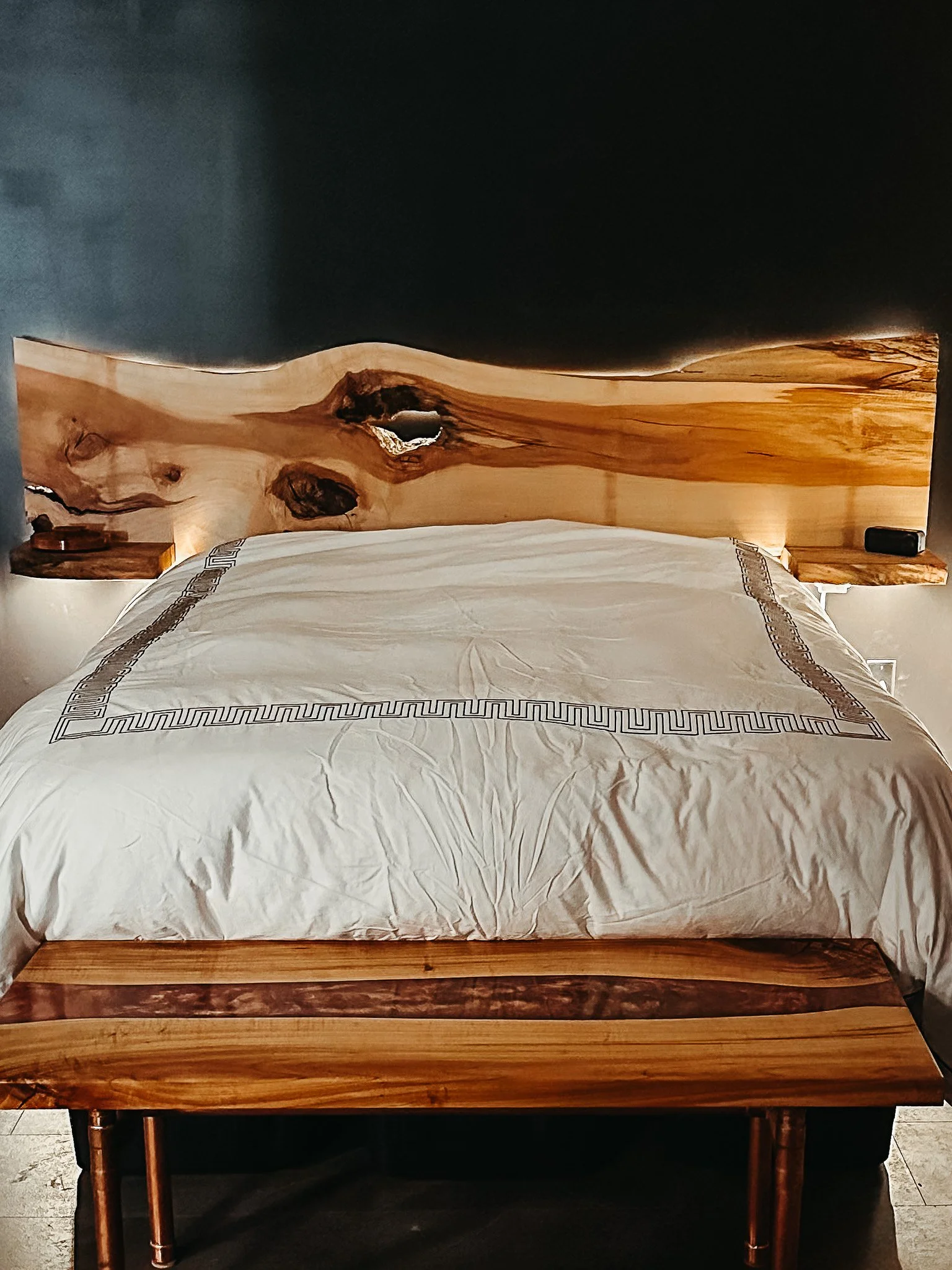 A wooden bed frame with a natural finish and a wood-paneled headboard, a white bedspread with embroidered trim, and two bedside tables with lamps.