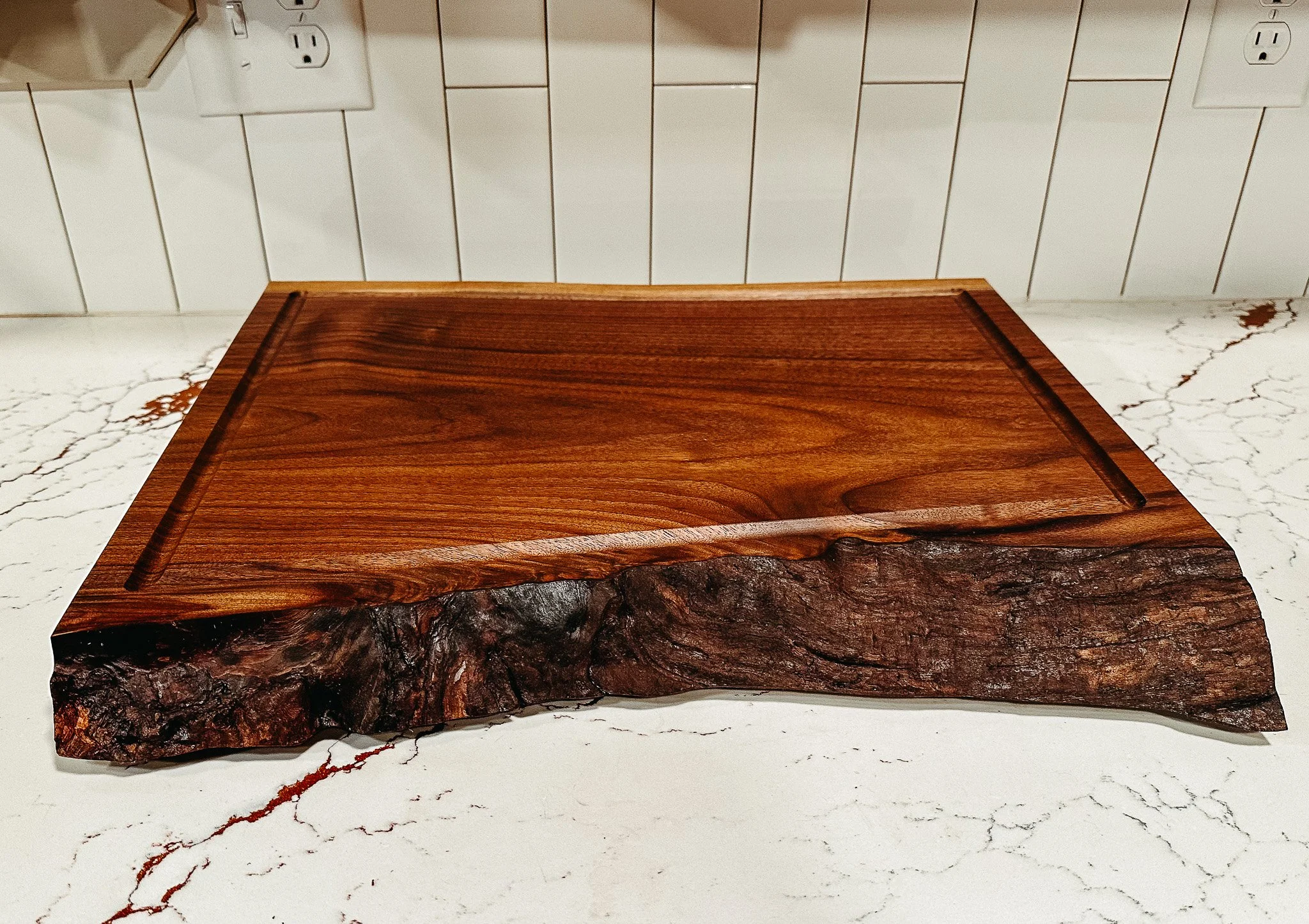 Close-up view of a wooden cutting board on a white marble countertop with electrical outlets and a white tiled wall in the background.