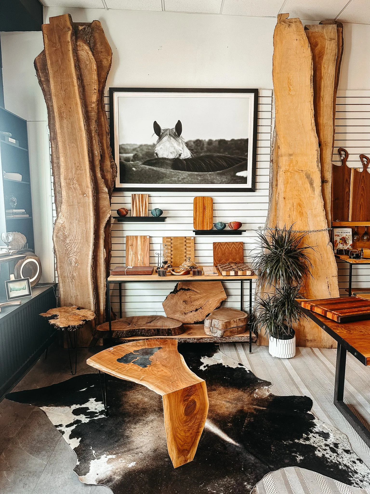 Interior of a decor shop showcasing rustic wooden furniture, including tables and stools, with a black and white horse photograph mounted on a wall behind the furniture, and a cowhide rug on the floor.