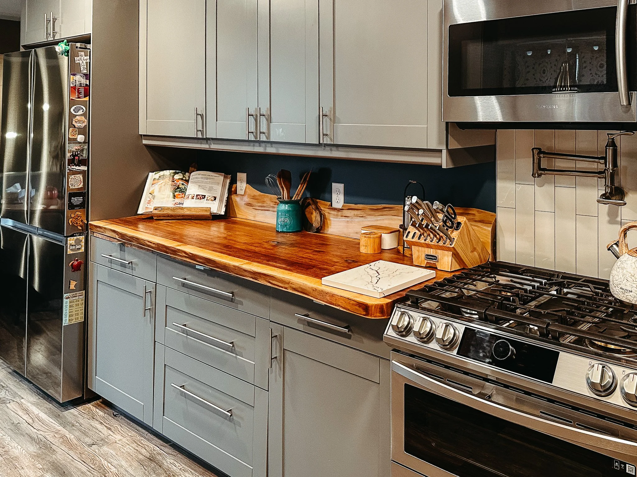 A modern kitchen with light gray cabinets, a wooden countertop, a stainless steel microwave, a gas stove, and a refrigerator with various stickers. There is an open cookbook, utensils in a teal container, and a cutting board on the countertop.