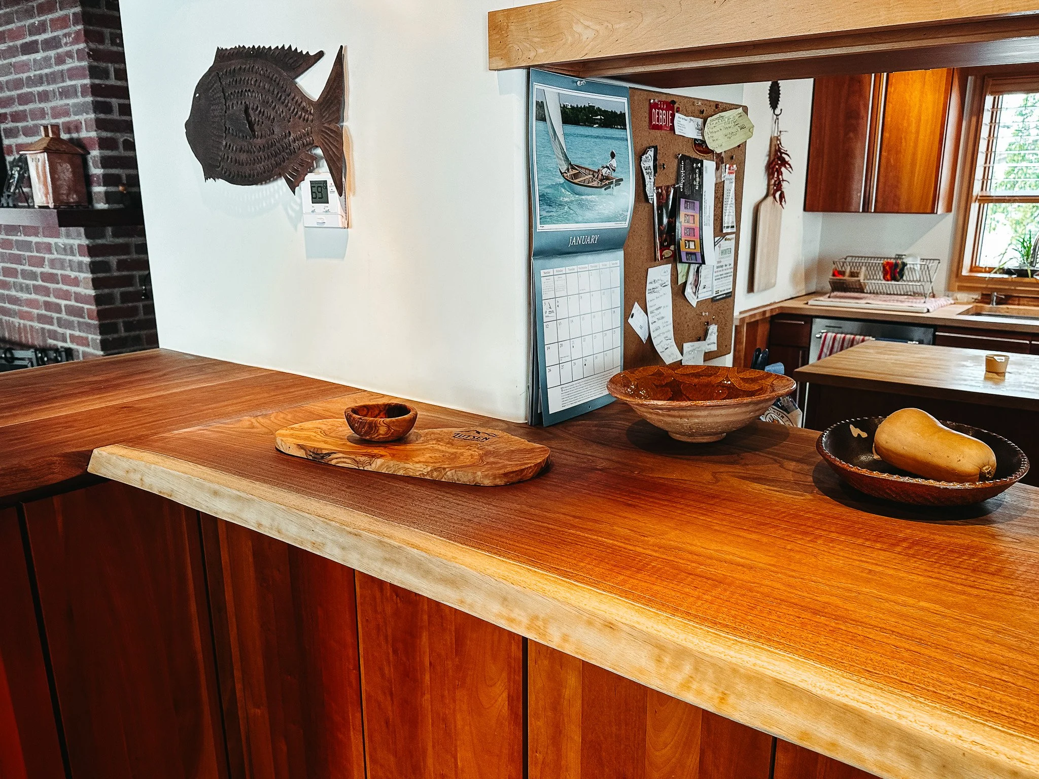 A wooden kitchen island with two different styles of wood counters, one darker and one lighter. On the island are a small wooden bowl, a large bowl with a yellow squash, and a small wooden dish. In the background, there is a white wall with a metal f