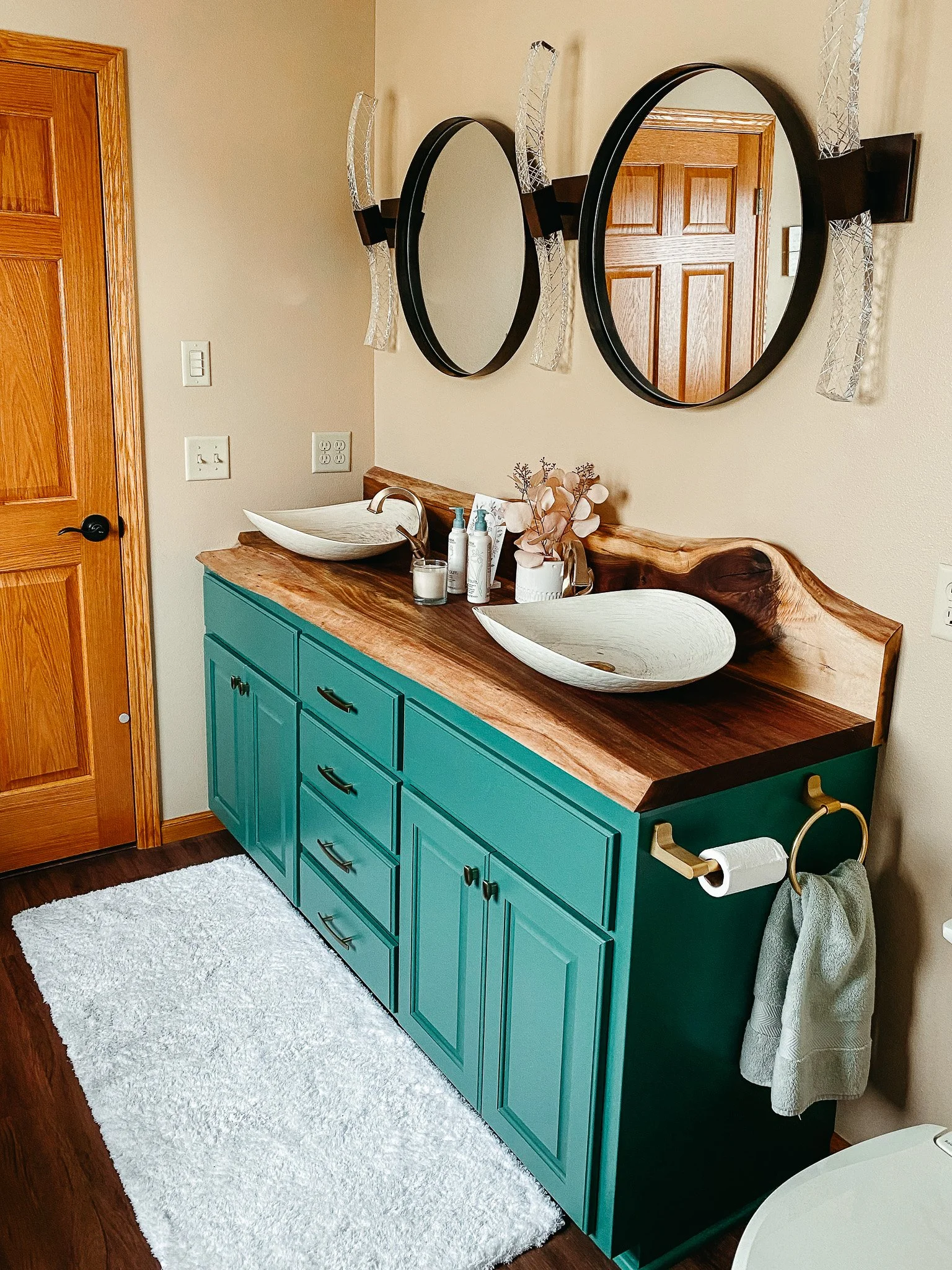 A modern bathroom vanity with a wooden countertop and two vessel sinks. The vanity is teal with multiple drawers and black handles. Above the sinks are two large round mirrors with black frames, mounted on a beige wall. To the right of the vanity, th