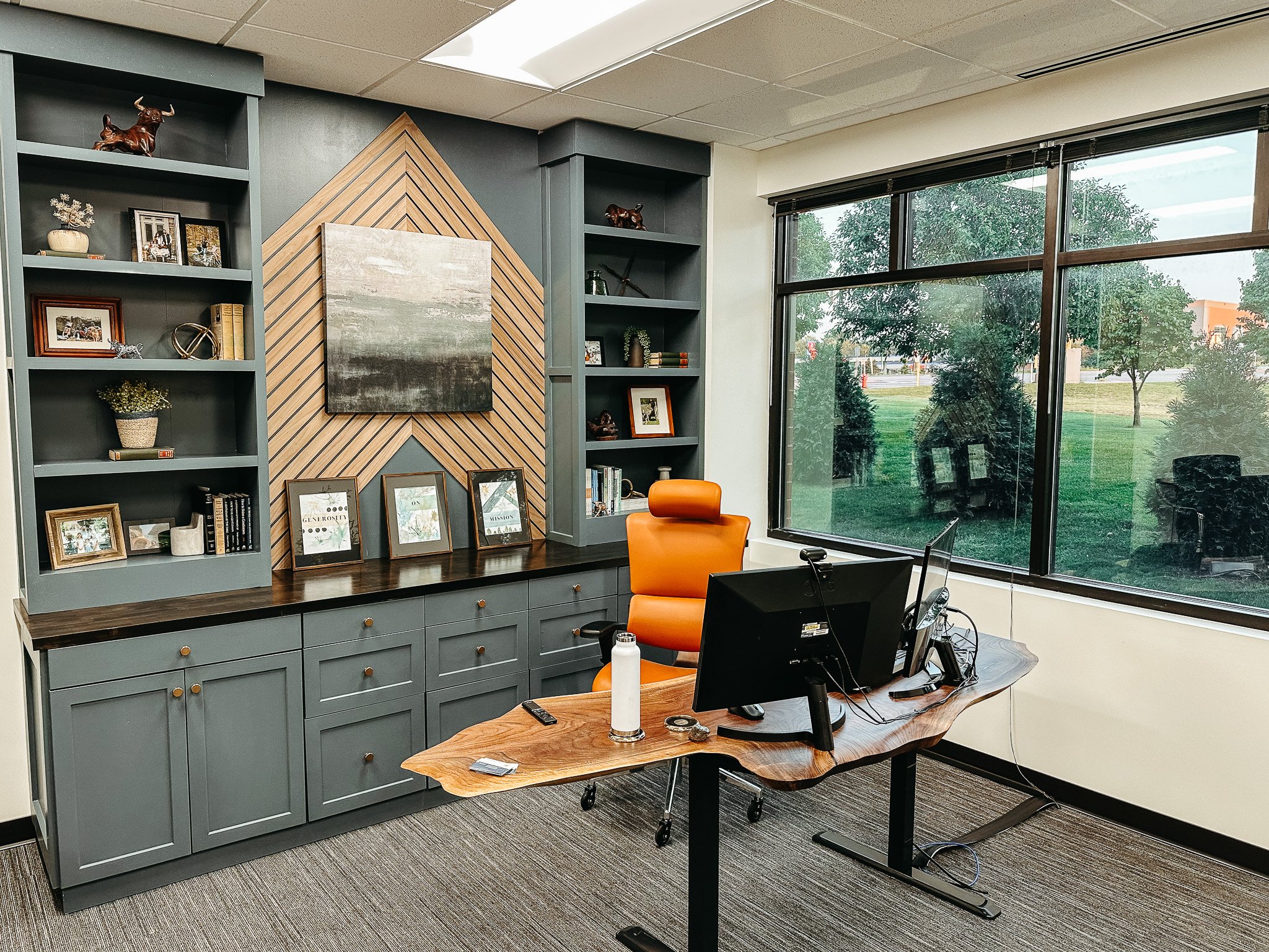 Office space with large windows, gray bookshelves with decorative items and picture frames, a wooden desk with a computer, and an orange office chair.