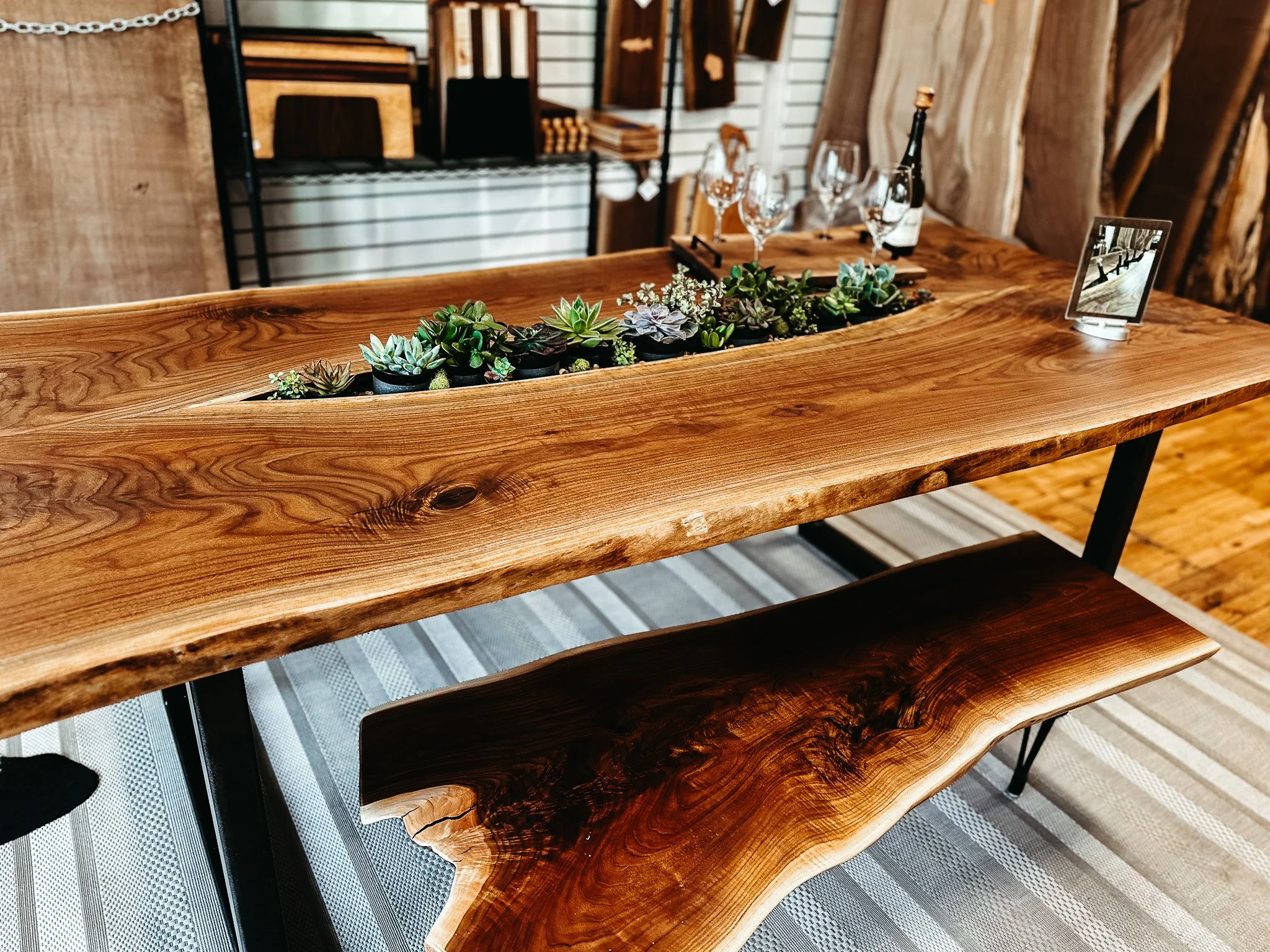 A wooden dining table with a natural, live-edge finish, decorated with a center arrangement of various potted succulents and small flowers, placed inside a long, narrow, rectangular wooden planting box. The table is set with four empty wine glasses, a tray with a bottle of wine, and a digital photo frame. In the background, there are shelves with wood pieces and a wall with wooden planks.