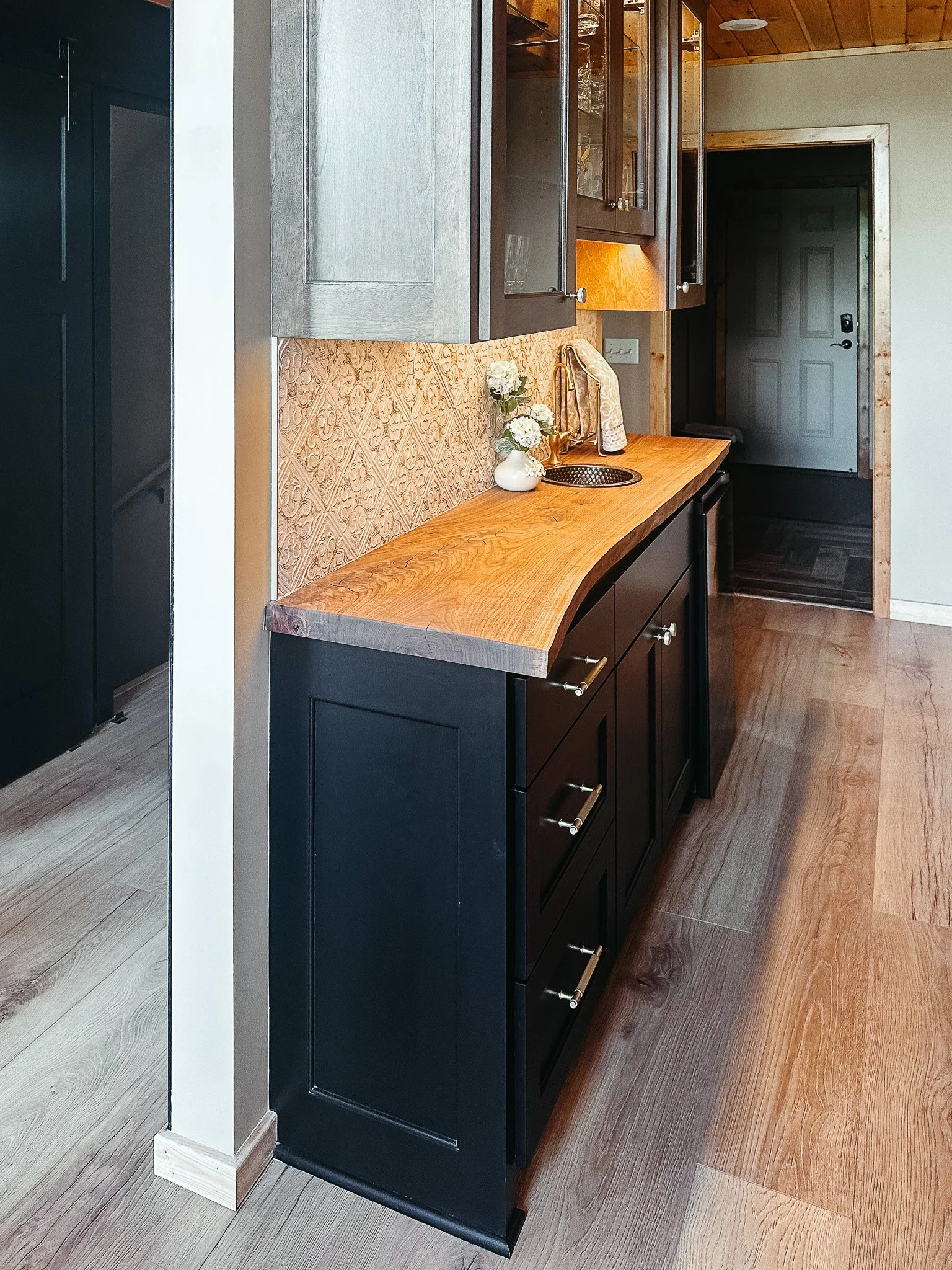 Kitchen counter with a wooden surface, a vase with flowers, a gold sink faucet, and a paper towel roll. Dark cabinets and some glassware are above, with a doorway at the back.