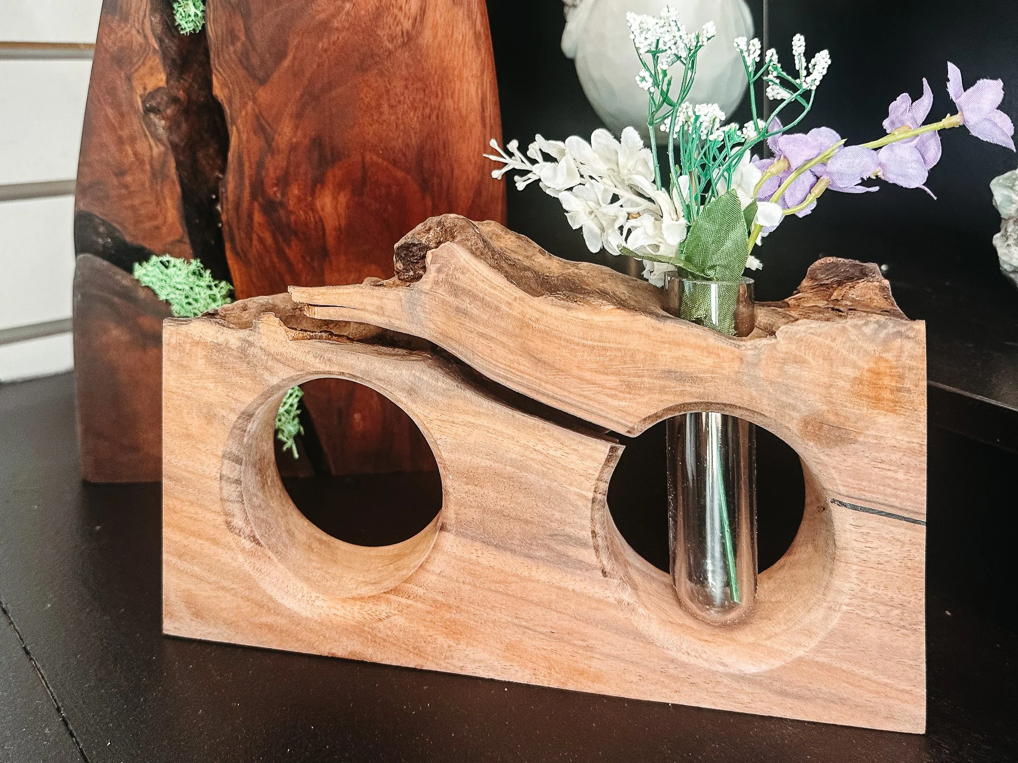 Decorative arrangement of artificial white and purple flowers in a small glass vase, positioned on a carved wooden stand.