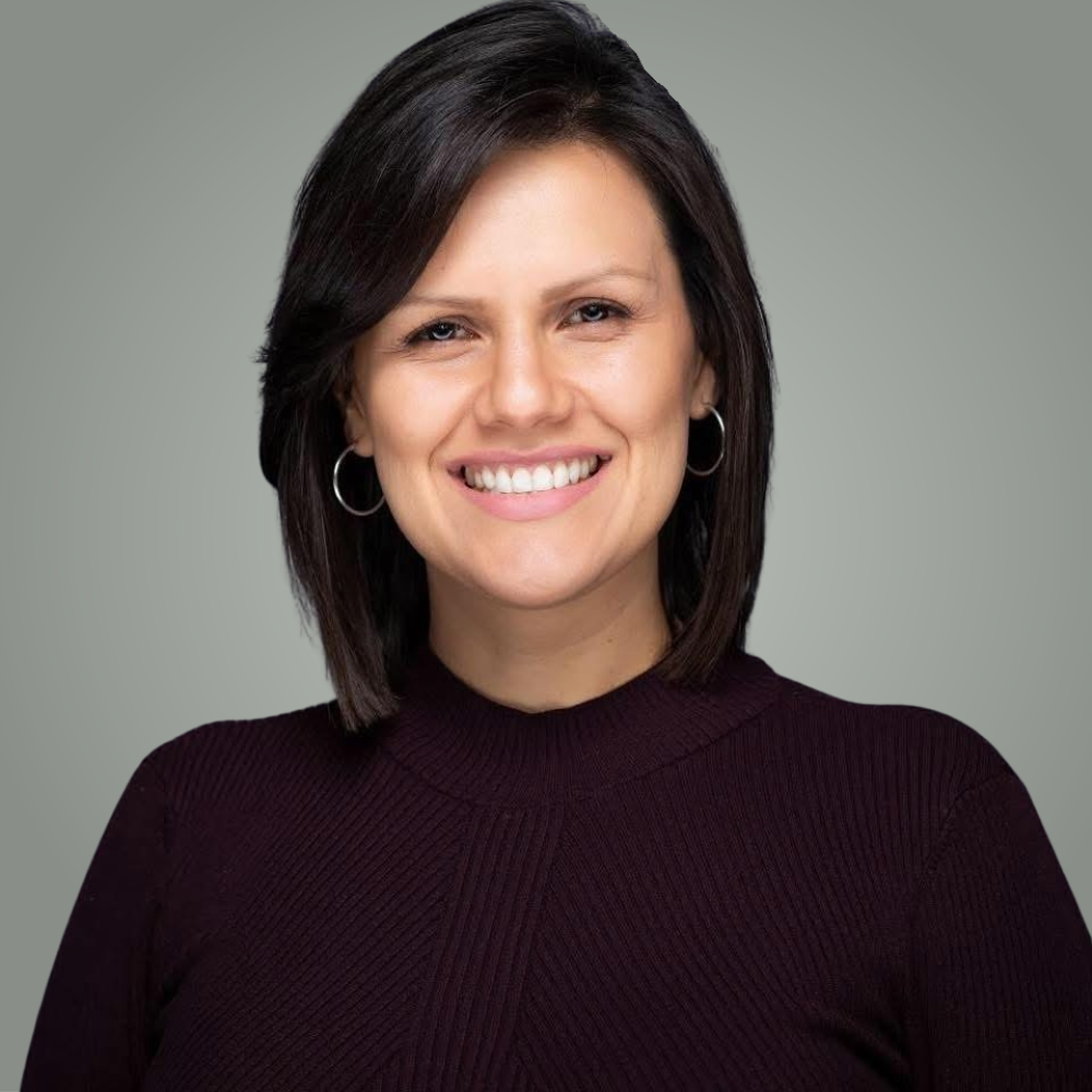 A woman with shoulder-length dark hair, wearing a dark long-sleeve top and hoop earrings, smiling against a plain gray background.
