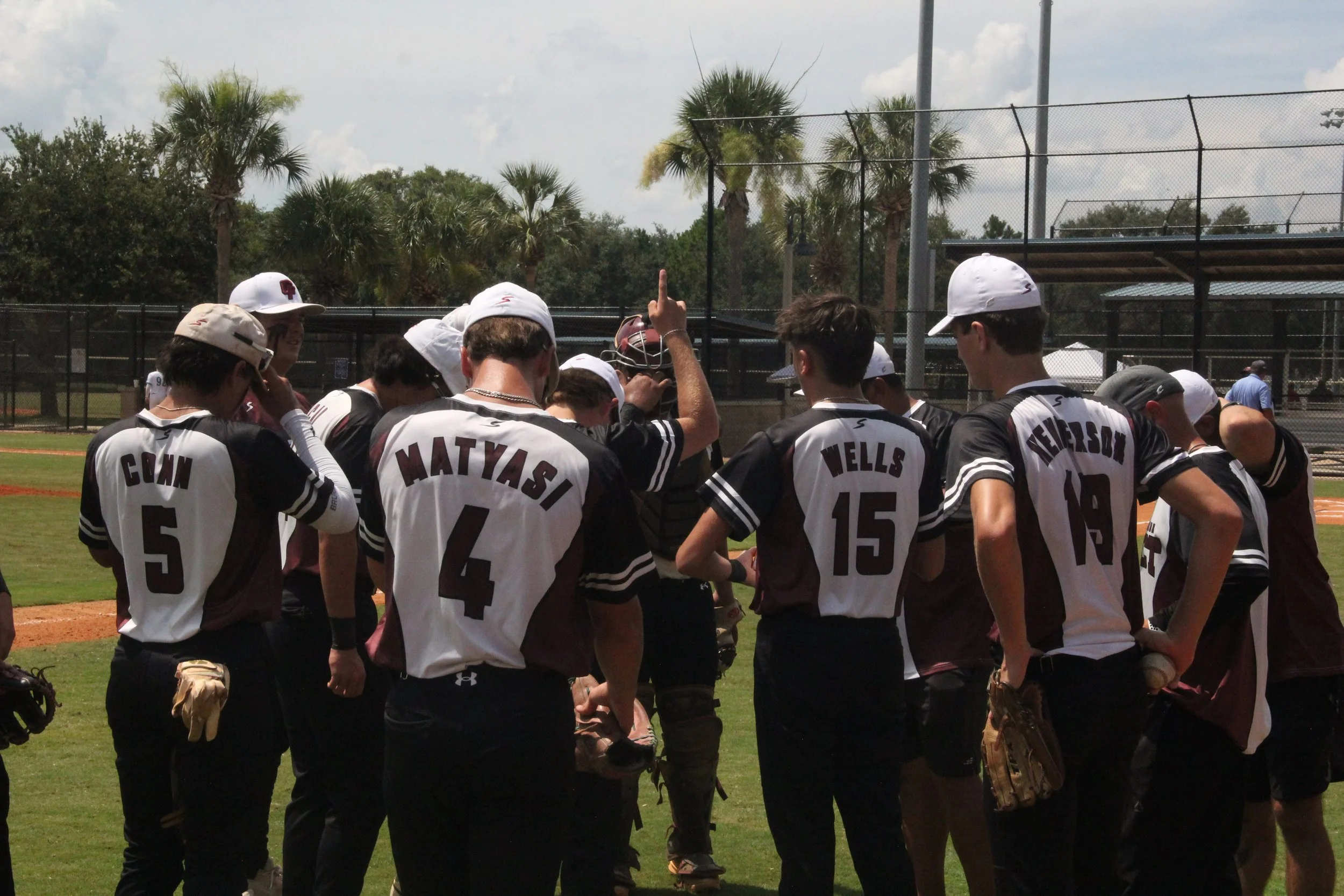 Baseball team huddling on the field during a game, with palm trees in the background.