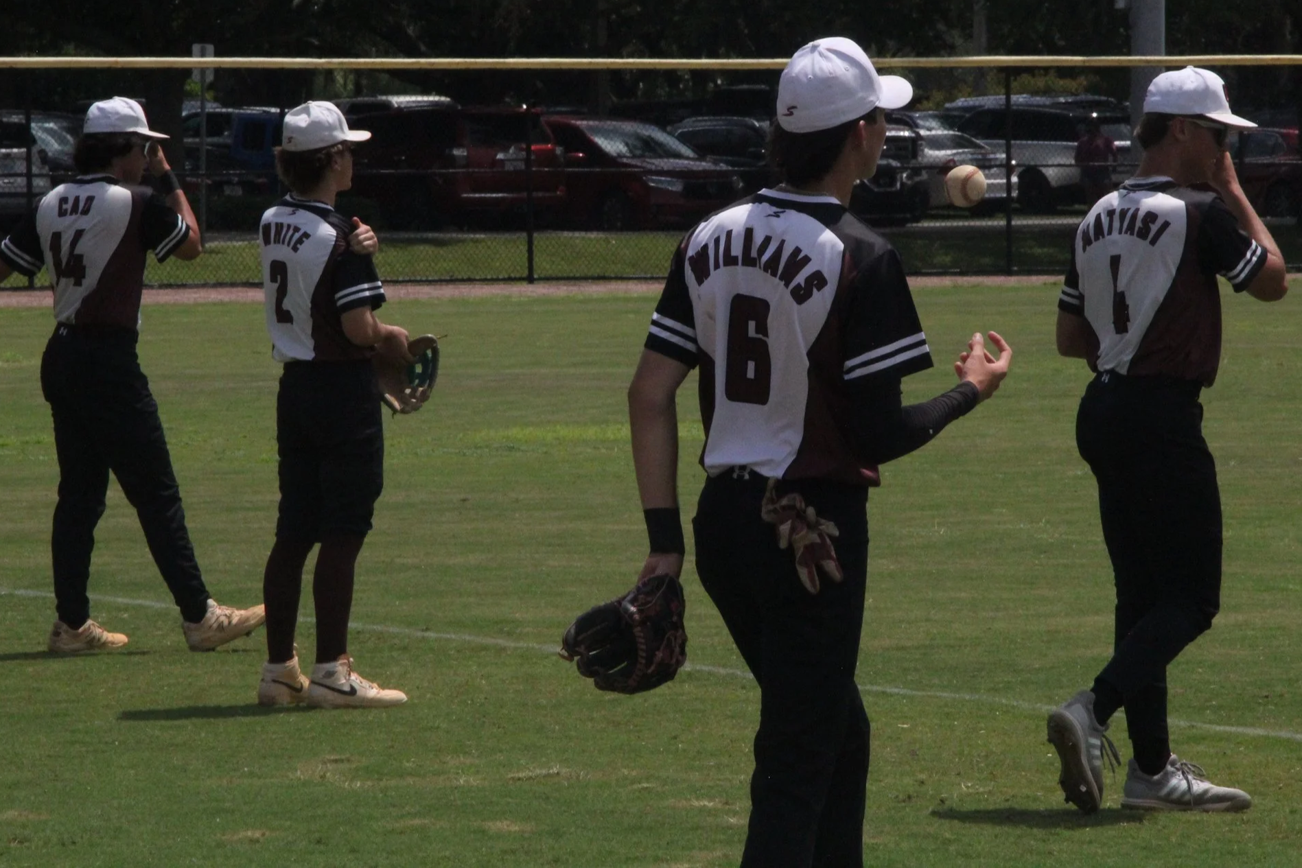 Young baseball players in matching uniforms and white caps on a field during daytime, preparing for a game, with parked cars and a chain-link fence in the background.