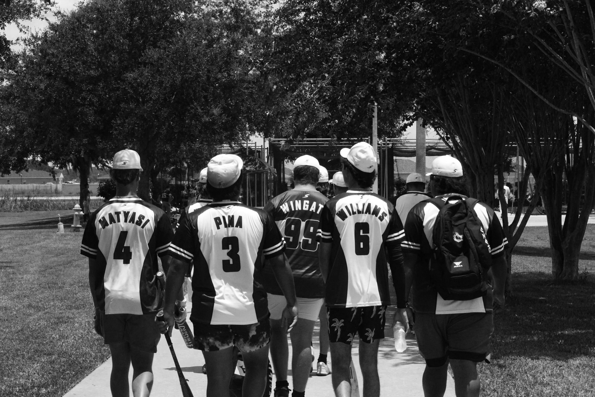 A group of players walking to batting cages, wearing jerseys and hats, some carrying backpacks and water bottles, with trees and a playground in the background.