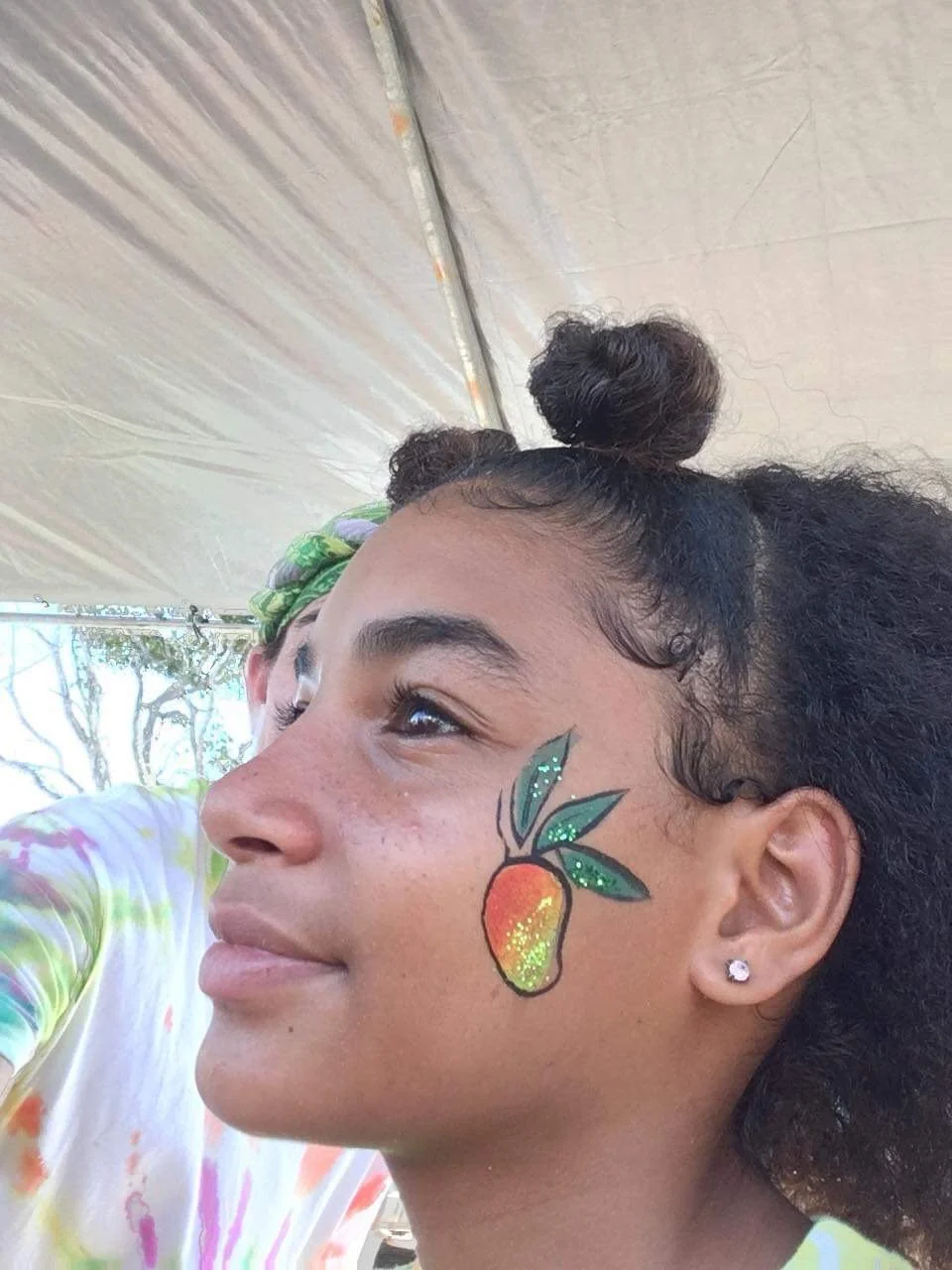 Young woman with face paint of a mango and leaves on her cheek, smiling and looking to the side, under a tent at an outdoor event.