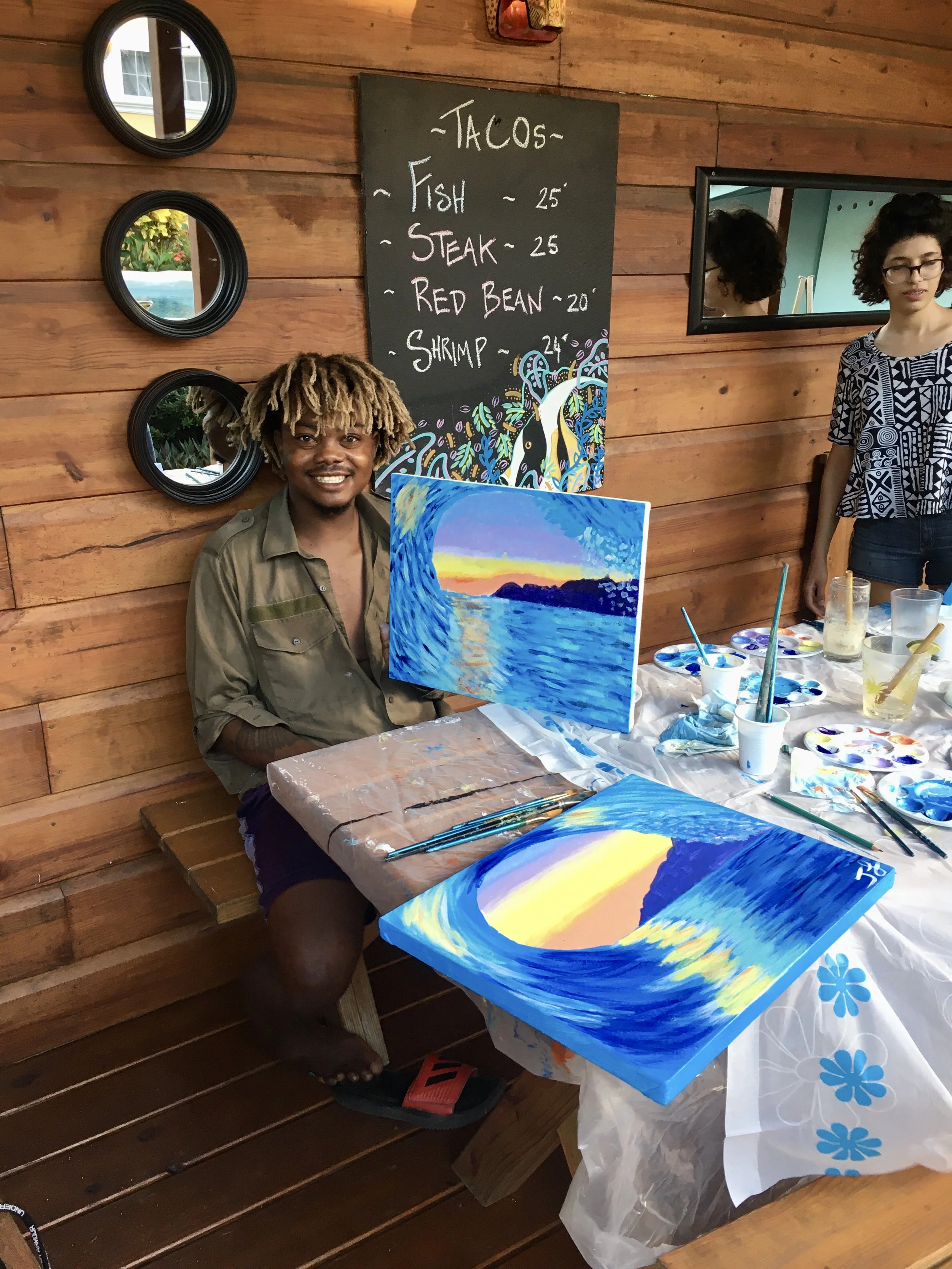 A person with dreadlocks smiling and holding a colorful painting of a sunset over water, seated at a table covered with painting supplies, in a wooden room with a chalkboard menu listing tacos and their prices.