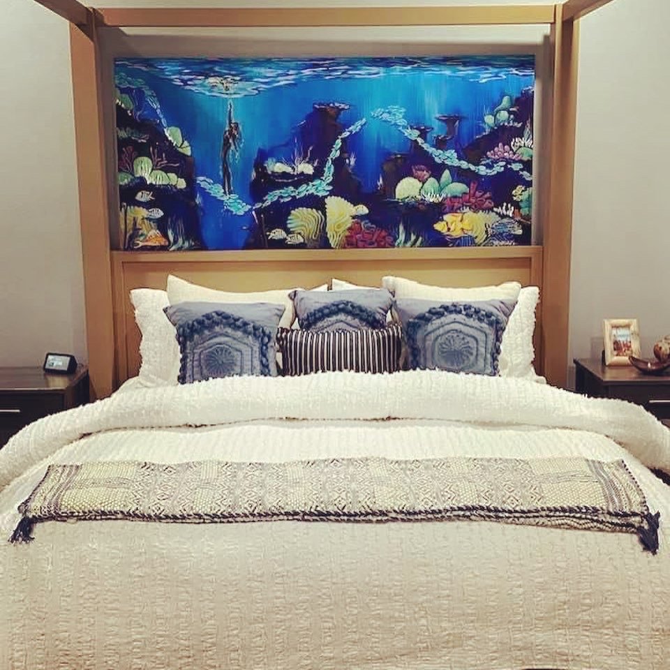 A bedroom with a large bed featuring multiple pillows and a textured white bedspread. Overhead, there's a painting of an underwater scene with a diver, coral, and rocks. On each side of the bed, there are bedside tables with framed photos and decorative items.