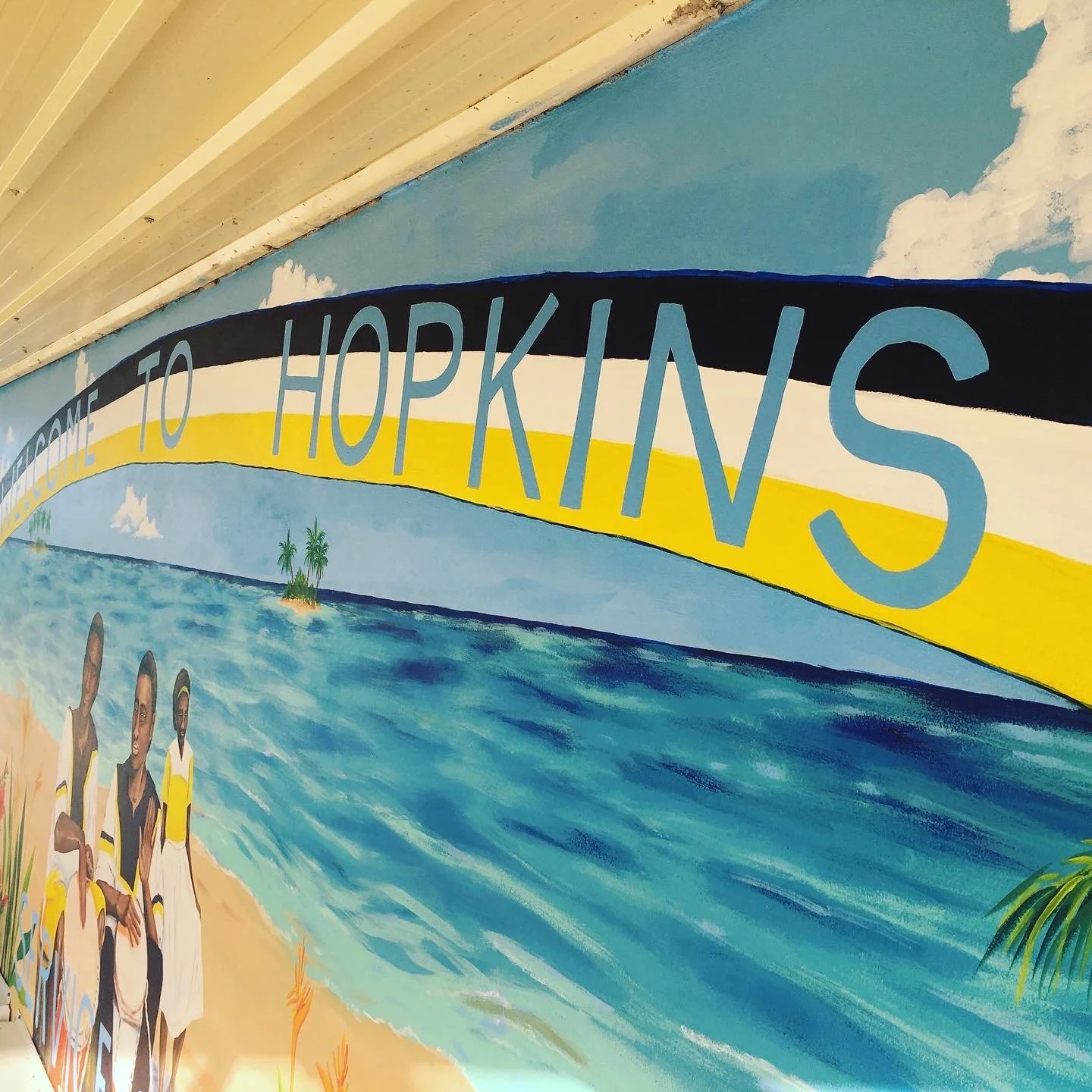 A colorful mural with the words 'Welcome to Hopkins' on a curved banner, depicting a tropical beach scene with palm trees, ocean waves, blue sky, and clouds, with images of people in beach attire.
