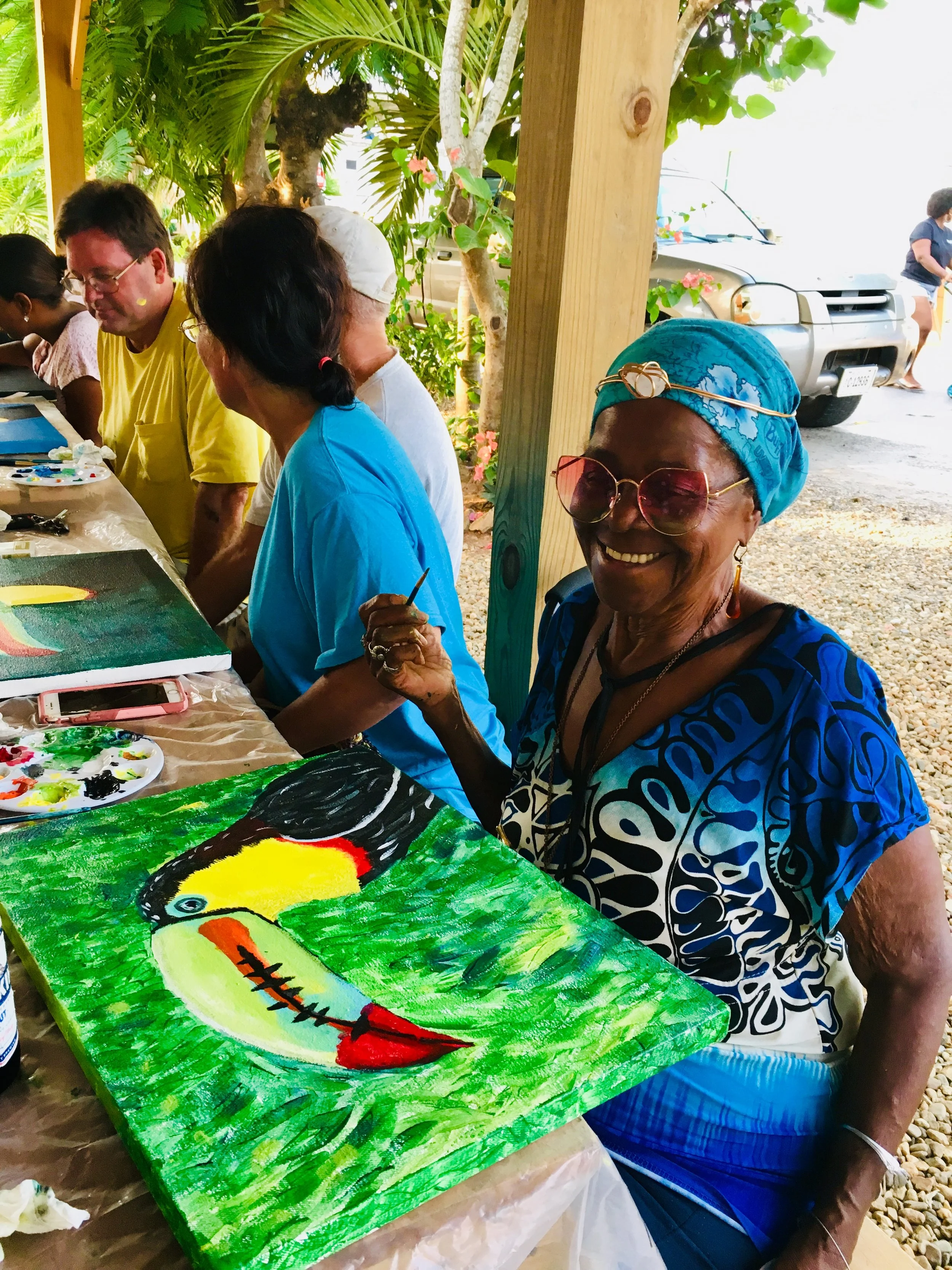 Smiling woman with blue headscarf and sunglasses showing a vibrant toucan painting at an outdoor art event with people at a table and parked cars in the background.