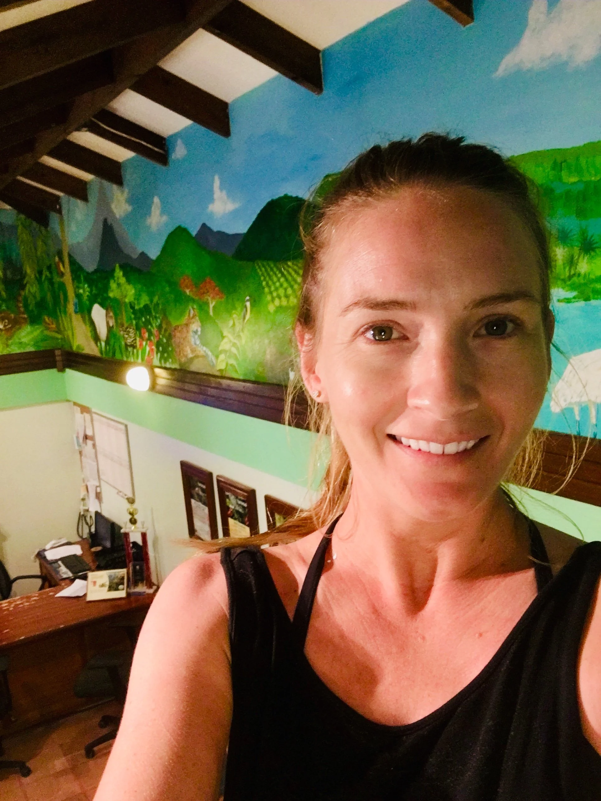 A woman with a big smile taking a selfie in a room with colorful wall art featuring a tropical landscape with mountains, clouds, trees, and a river. There is a wooden desk with assorted papers and books behind her.