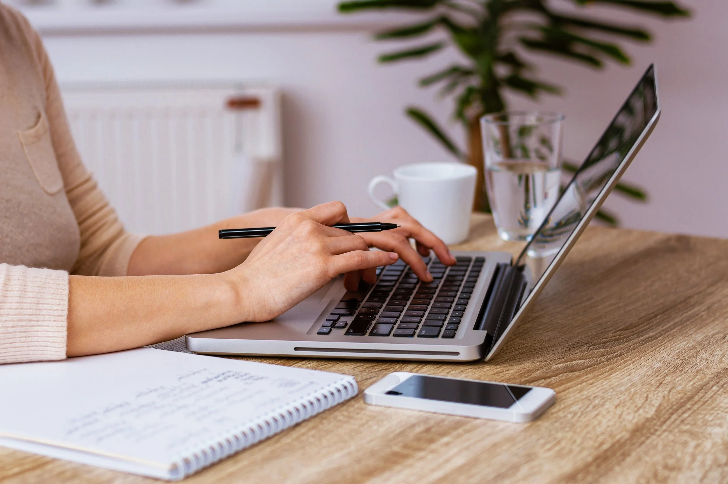 Person working on a laptop at a wooden desk with a smartphone, notebook, pen, coffee mug, and glass of water nearby.