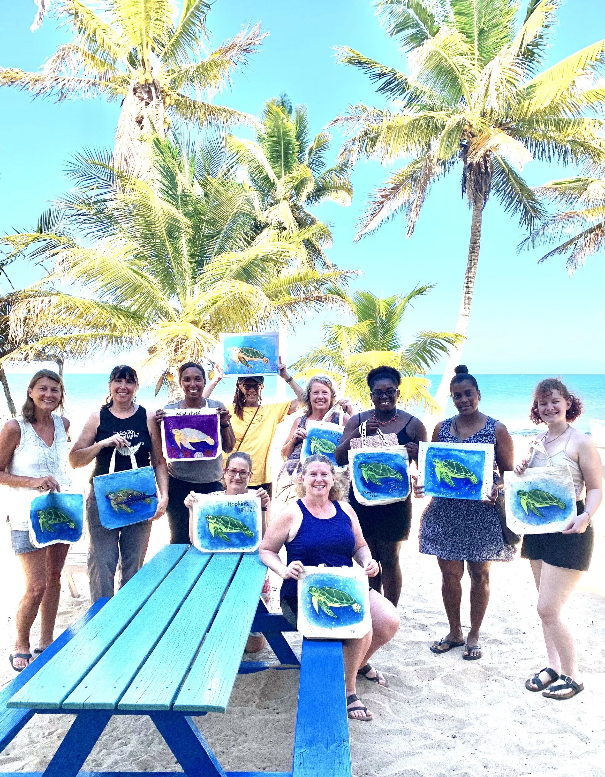 Group of people at the beach holding up paintings of turtles, with palm trees and a blue sky in the background.