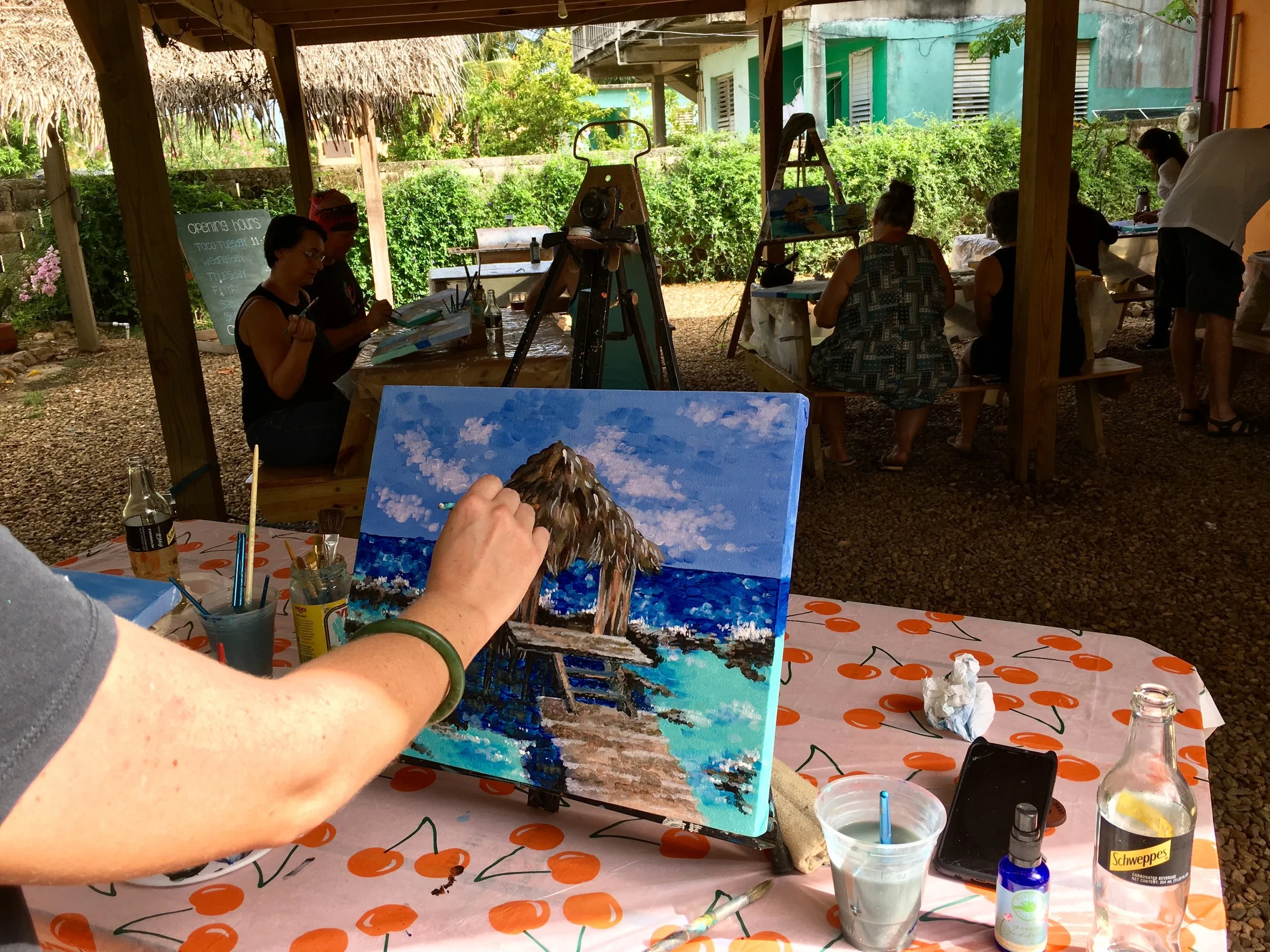 People painting landscape artwork outdoors under a wooden structure with green foliage and buildings in the background. Painting supplies are on a table with a cherry pattern tablecloth.