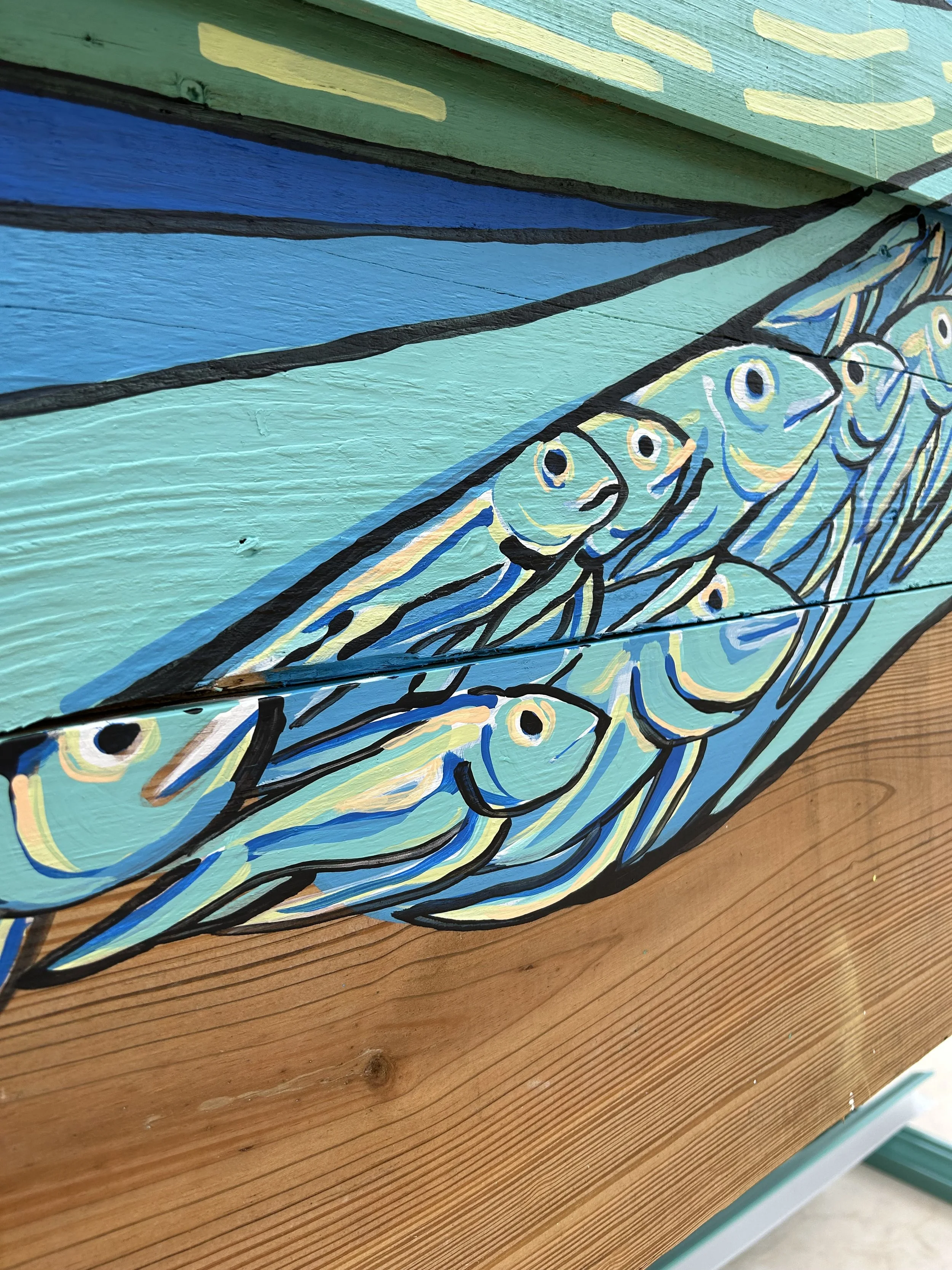 Close-up of a colorful wooden fish mural on a wooden surface, featuring blue, yellow, green, and black outlines.