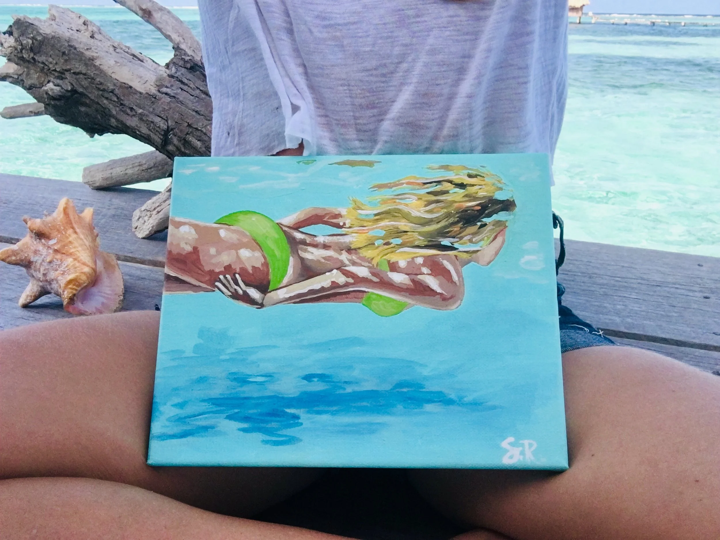 A hand-painted picture on canvas depicting a matcha latte with green lime, a sea shell, and orange slices on a blue background. The card rests on a person's legs at a beach with clear turquoise water and a wooden railing.