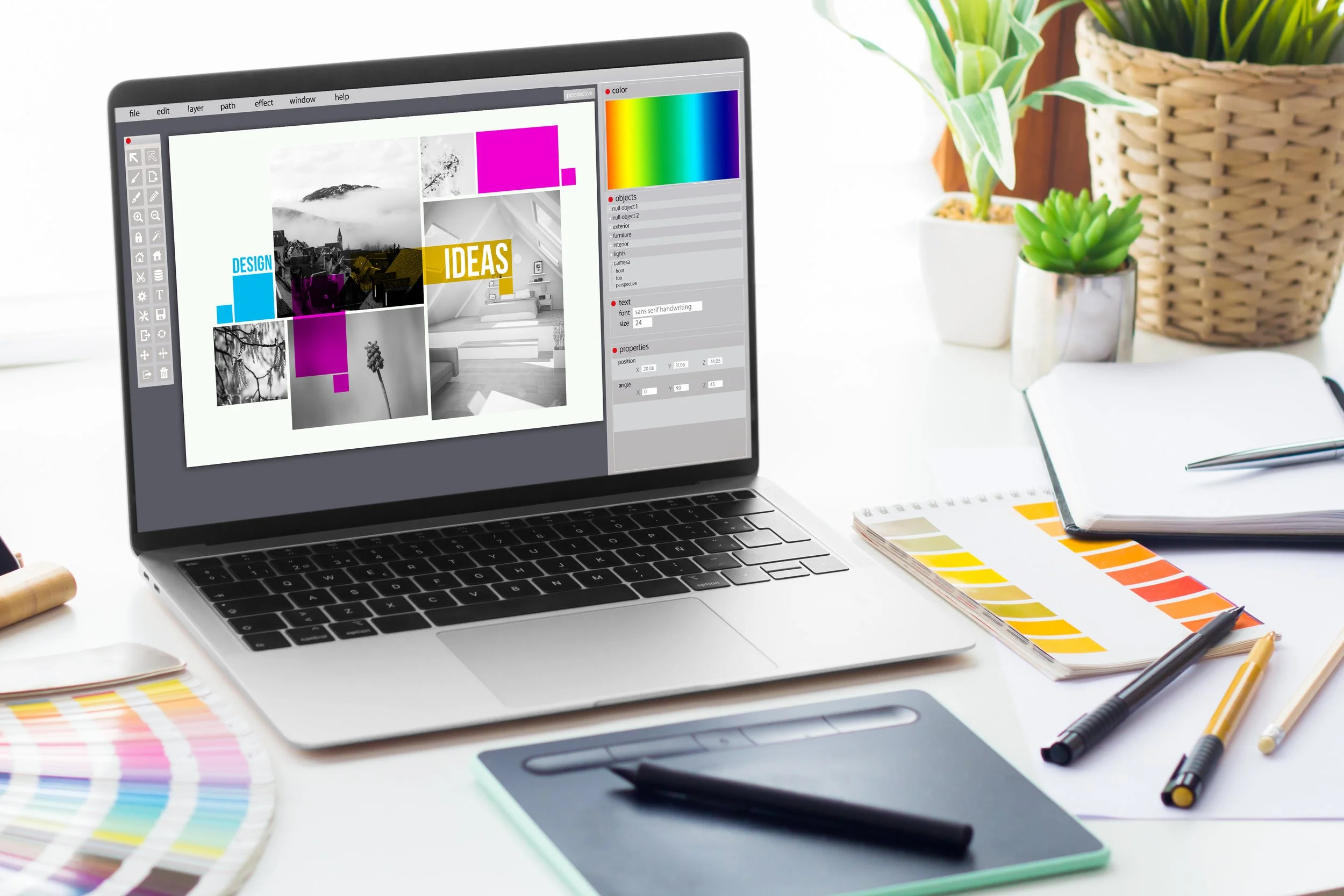 Open laptop displaying design software with images, color palette, and text editing tools on a white desk with color swatches, notebooks, pens, and potted plants nearby.