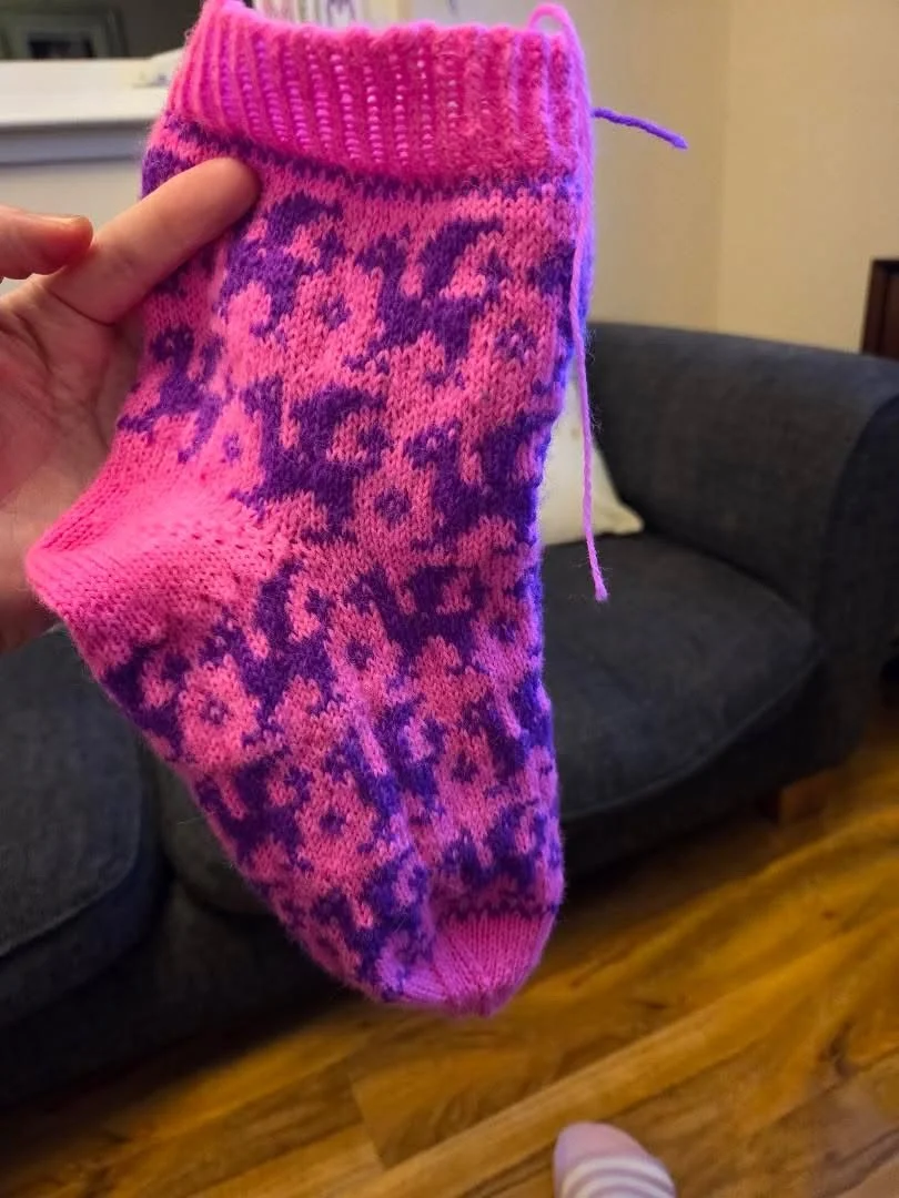 A person's hand holding a pink and purple knitted sock with a dragon pattern, in front of a dark gray couch on a wooden floor.