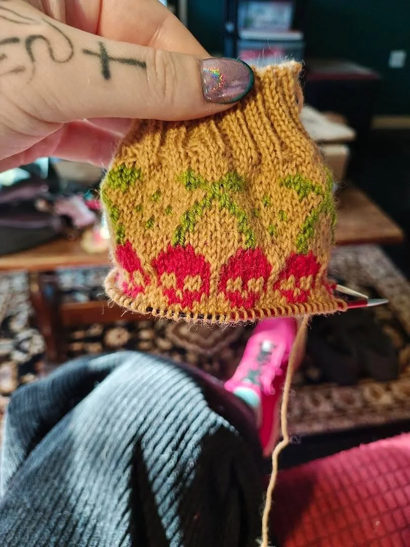 Close-up of a small, yellow-orange knitted sock with red and green devil-horned skull berry pattern, held by a hand with a colorful, glittery nail. The background shows a living room with a blurred couch and a television.