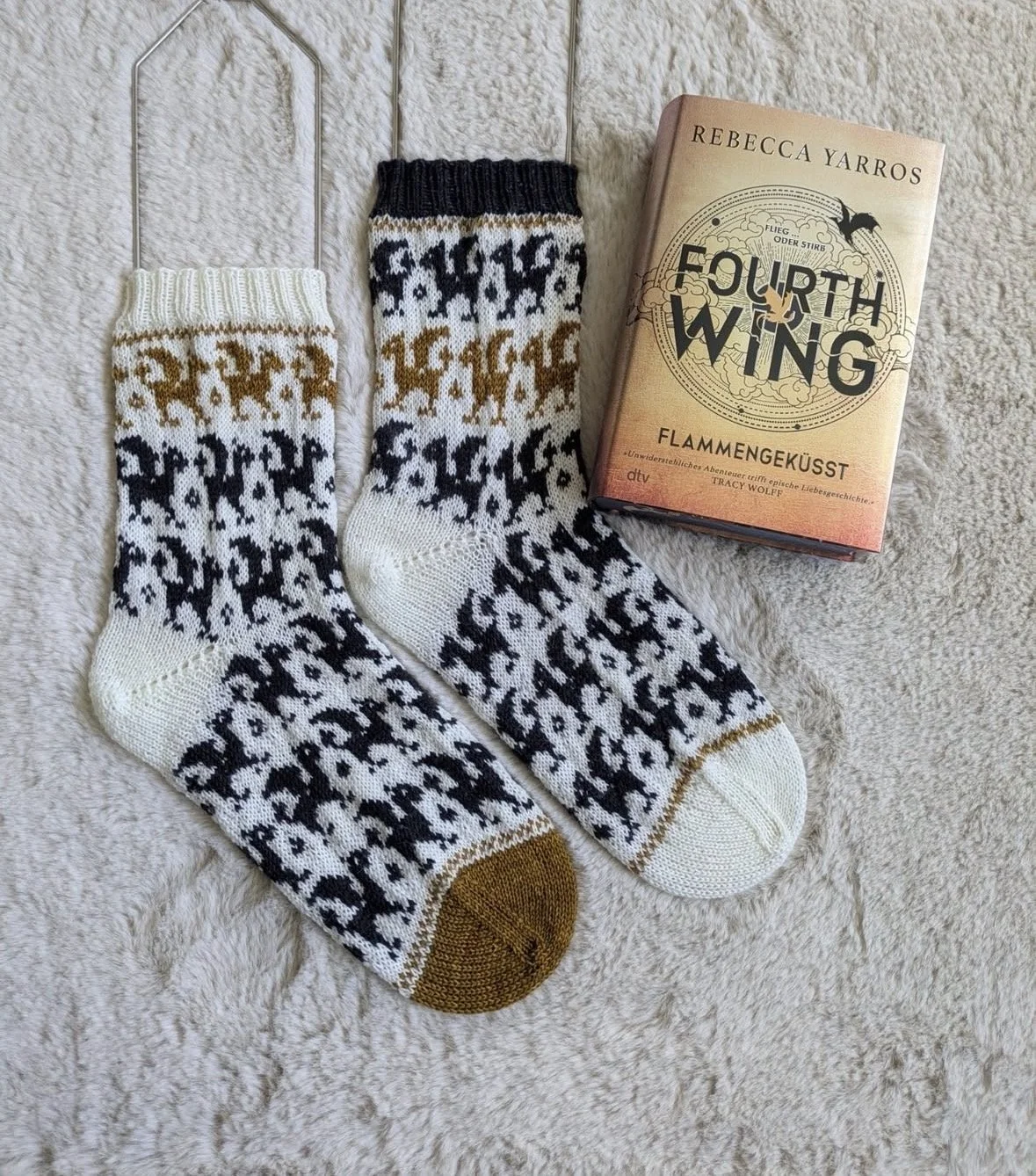 Pair of knitted dragon socks with a black, white, and gold dragon pattern and a book titled "Fourth Wing" by Rebecca Yarros, placed on a beige, furry surface.