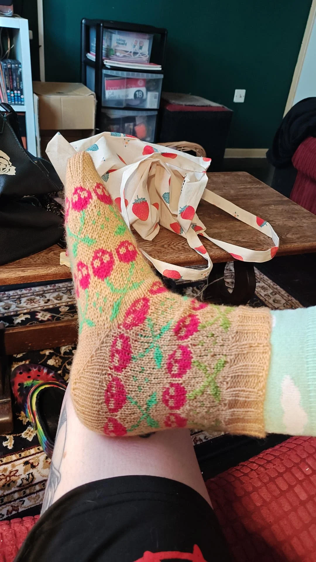 Person sitting with legs crossed wearing socks with a pink and green cherry skull pattern, on a black shirt, on a red cushion, in a room with a wooden table, a black bag, a strawberry-patterned tote bag, a black shelf, and a green wall.