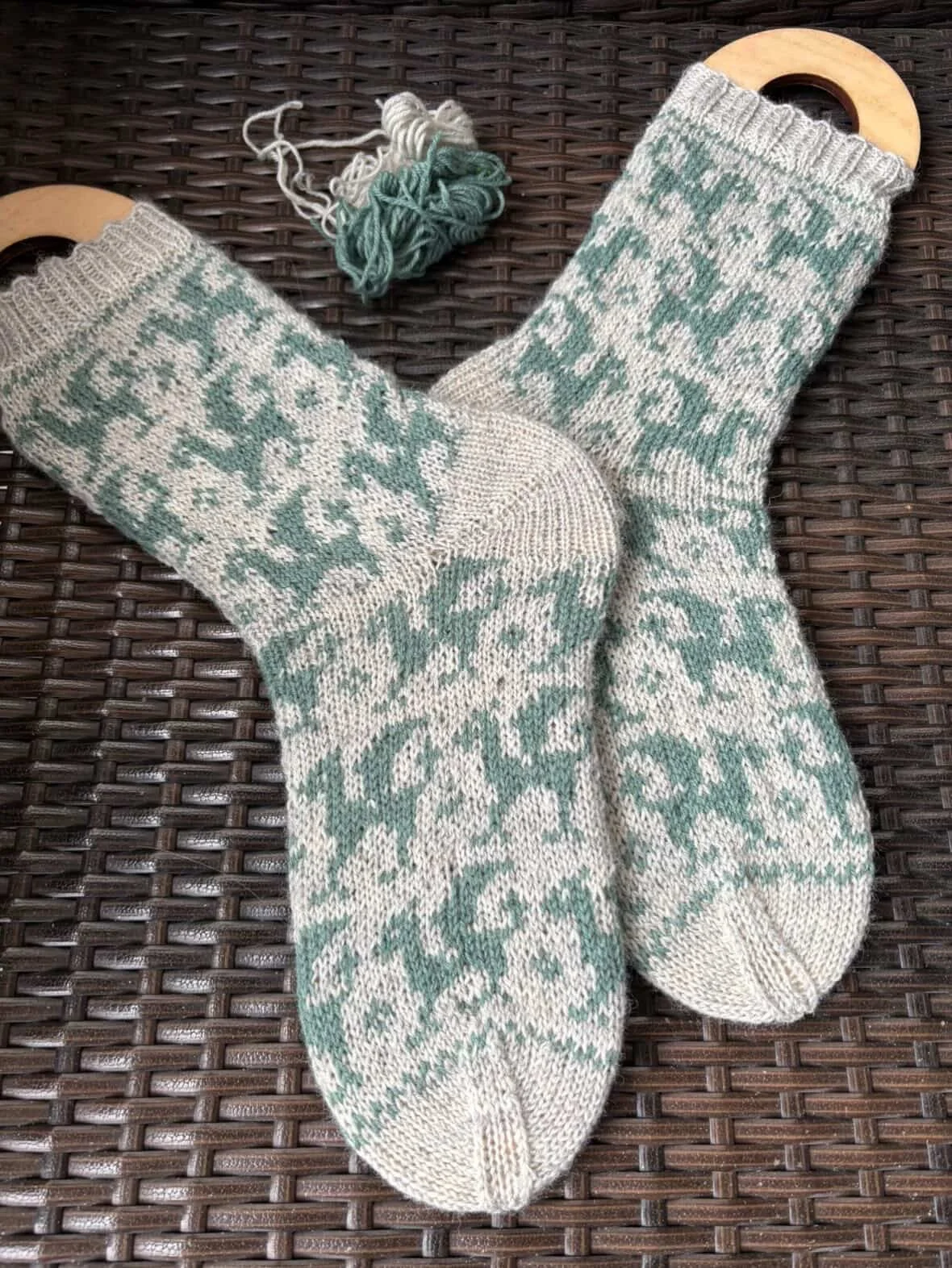 A pair of knitted socks with a dragon pattern, hanging on a dark woven surface with a small ball of yarn nearby.