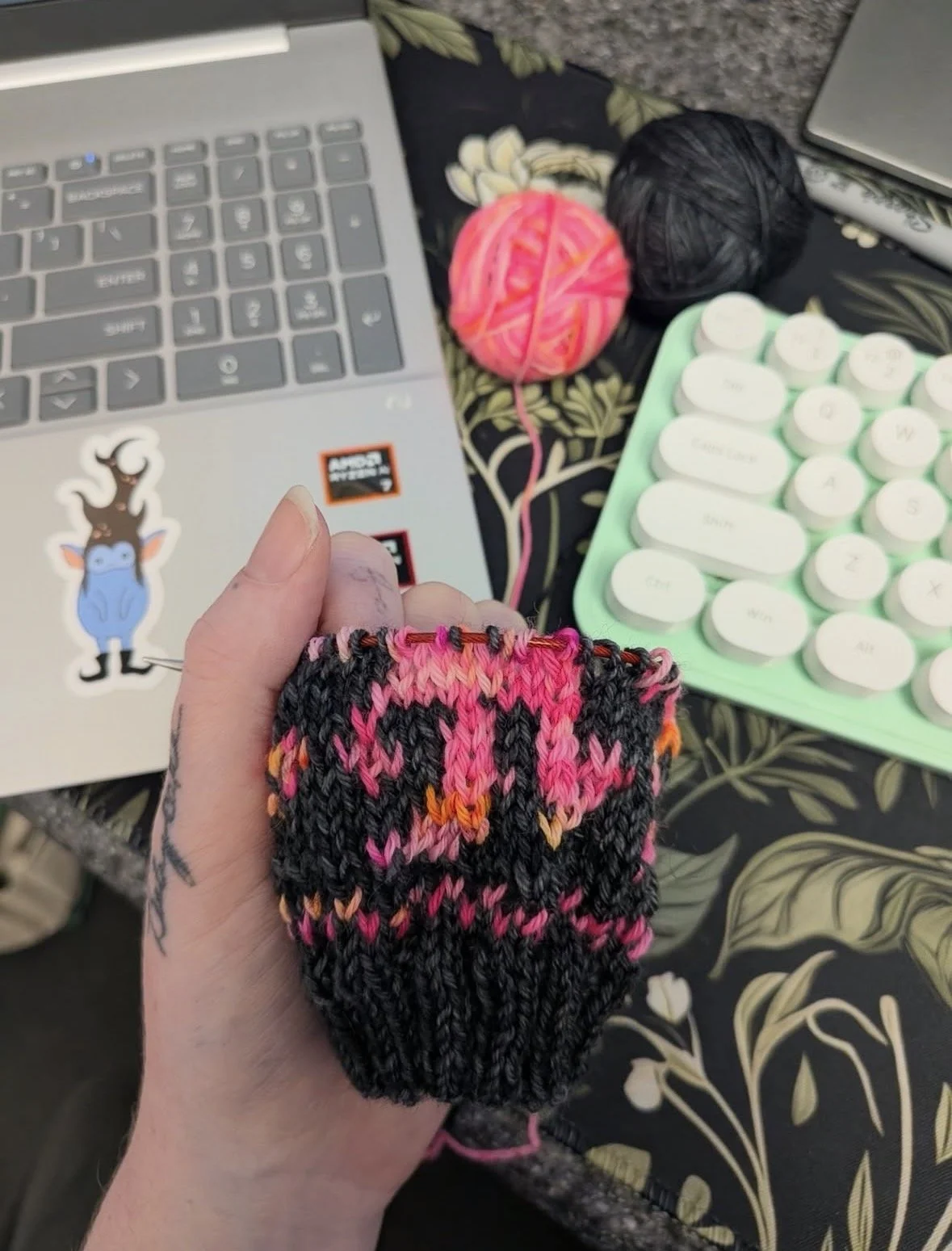 A person's hand holding a colorful knitted sock with black, pink, and orange yarns. On a table, there are two balls of yarn in pink and black, a laptop with a sticker of a blue cartoon character with horns, a small keyboard, and a floral-patterned fa