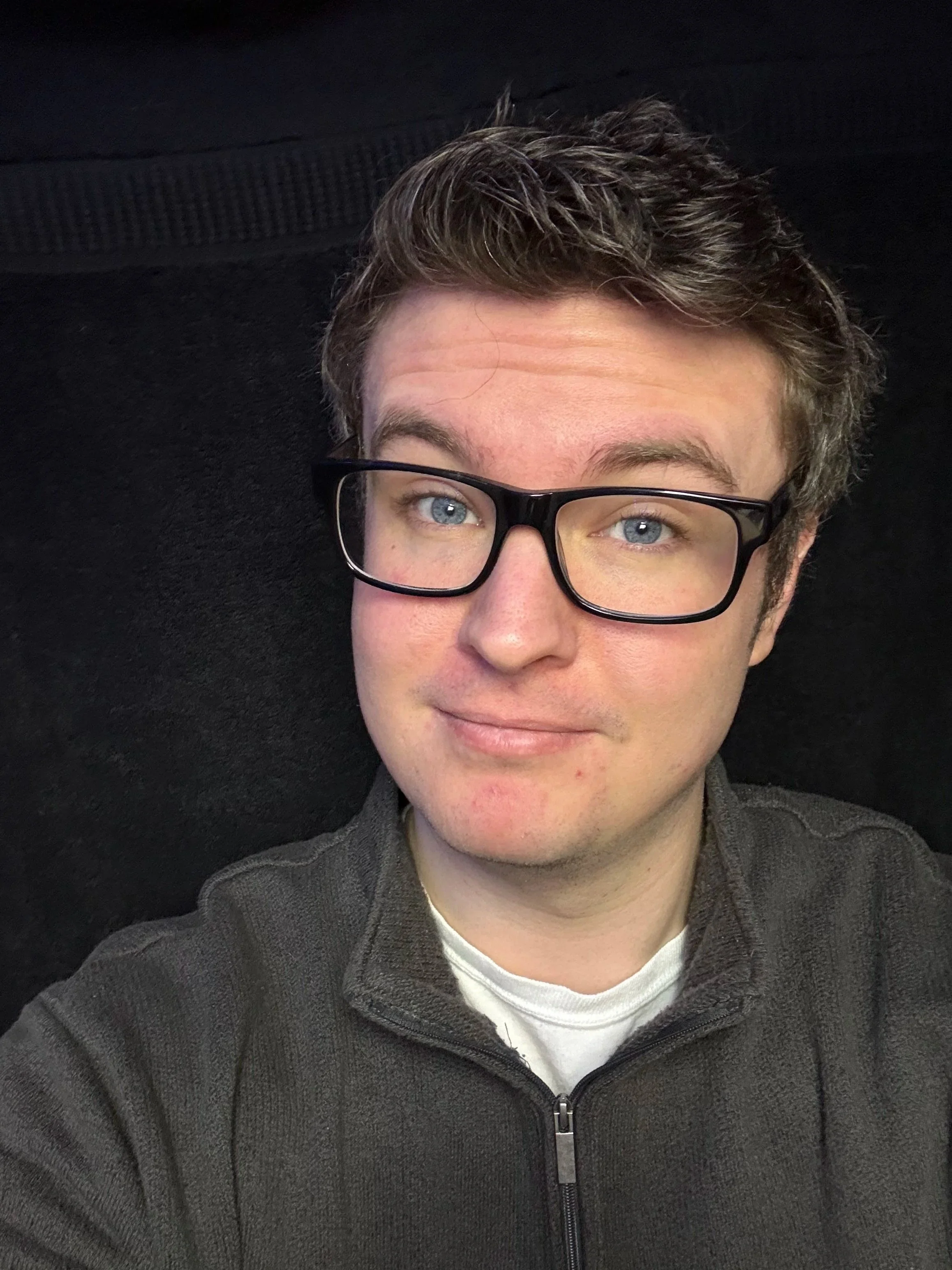 A young man with light skin, short brown hair, and blue eyes wearing black glasses and a dark zip-up sweater, smiling slightly, against a black background.