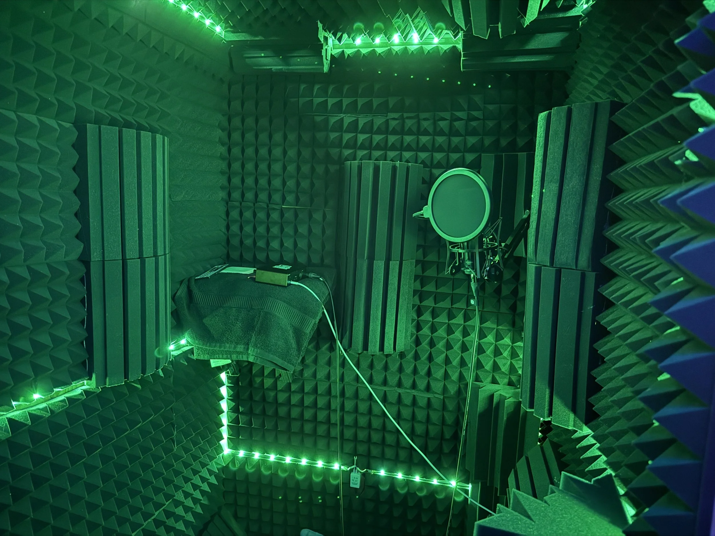 A soundproof recording booth with green LED lighting, acoustic foam panels on the walls and ceiling, a microphone with pop filter on a stand, and a small table covered with a cloth holding audio equipment.