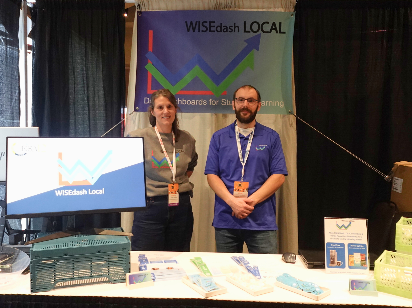 Two people standing behind a booth at a conference, with a large banner displaying the logo 'Winsights' and graphs. The booth has a computer monitor, brochures, and a sign about prize chances.