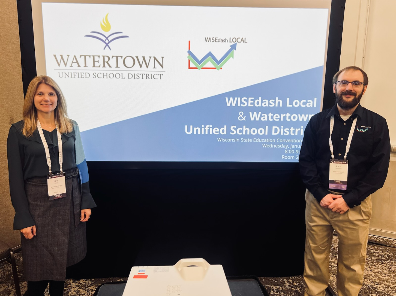 Two people standing in front of a presentation screen displaying consortium information while attending a Wisconsin State Education Convention.