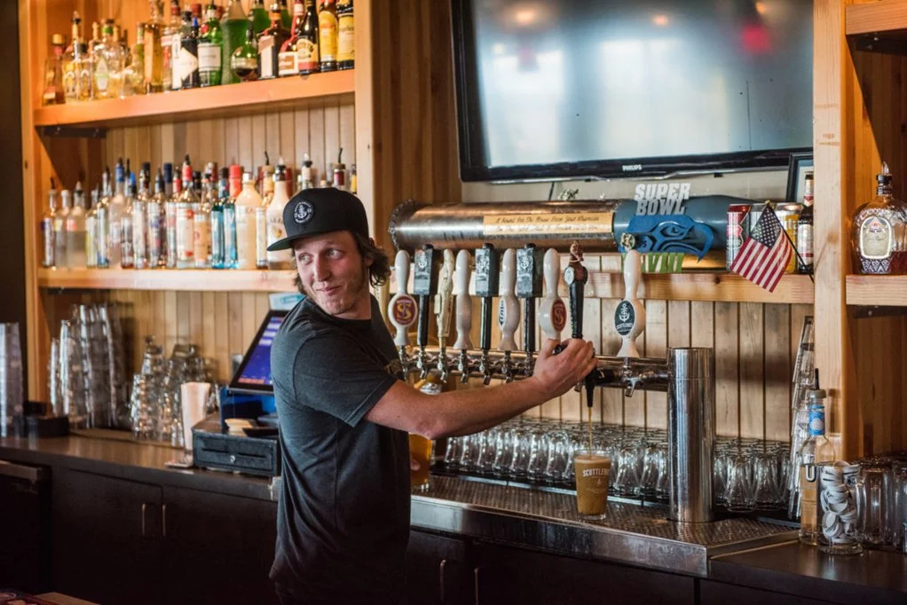 A bartender pouring draft beer at a bar with wooden shelves filled with bottles of alcohol, a large television, and American flags.