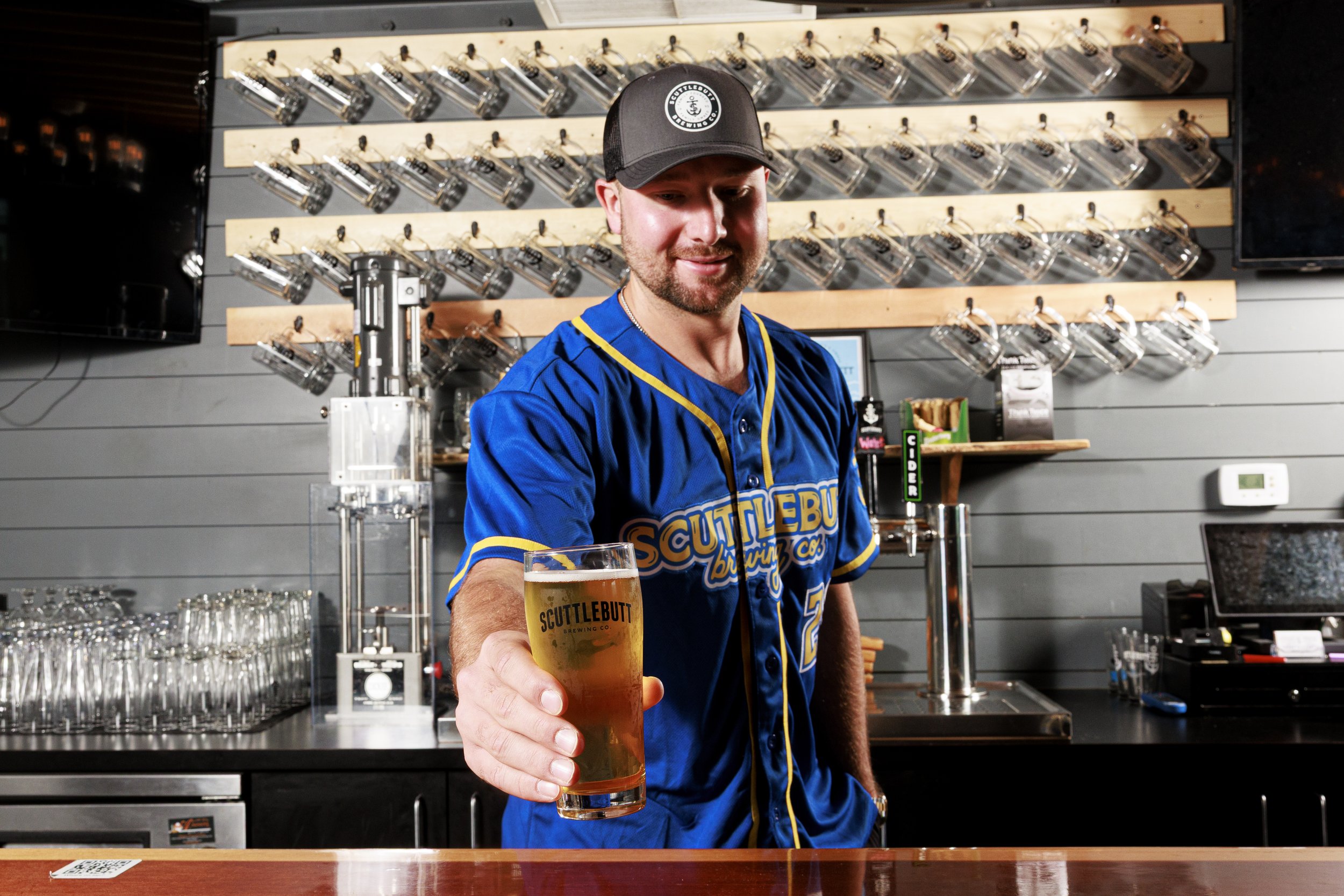 Cal Raleigh in a blue sports jersey and black cap serving a glass of beer in a bar with multiple beer taps on a wall behind him.